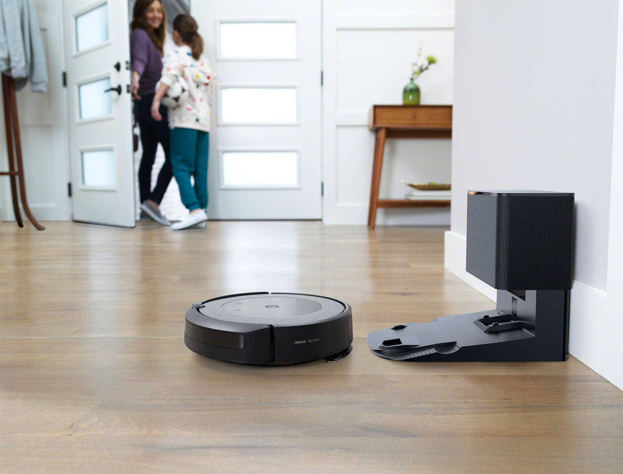 Ein schwarzer Roboter-Staubsauger und eine Ladestation auf einem Holzboden. Eine Tür mit zwei Personen im Hintergrund.