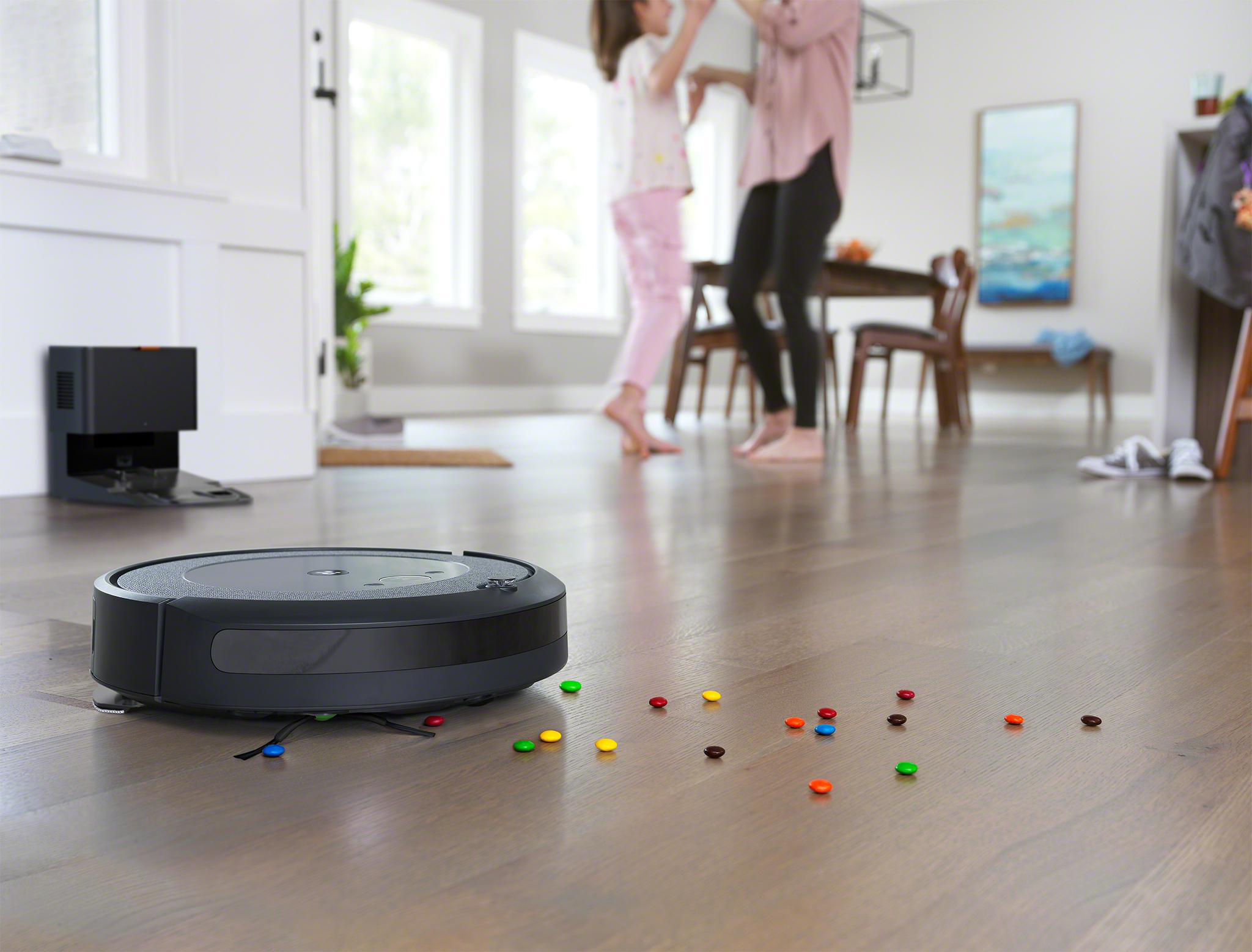 Ein schwarzer Roboterstaubsauger steht auf dem Boden. Bunte Süßigkeiten verstreut. Zwei Personen tanzen im Hintergrund.