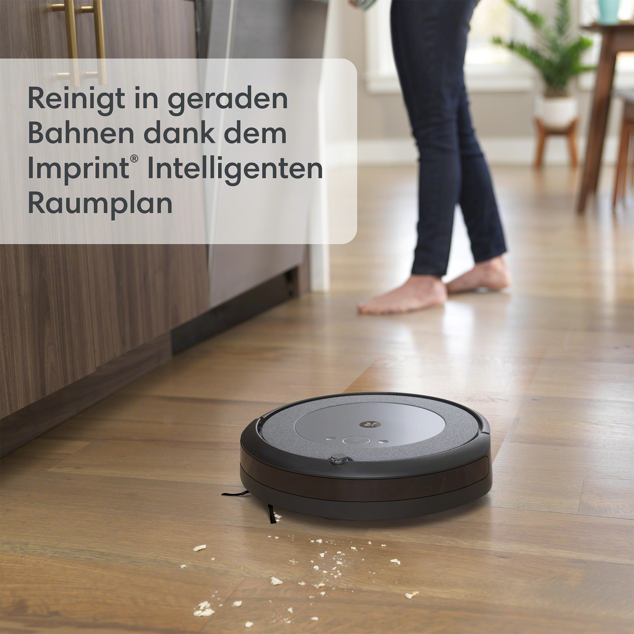 Ein schwarzer Roboter-Staubsauger reinigt einen Holzboden. Im Hintergrund steht eine Person.