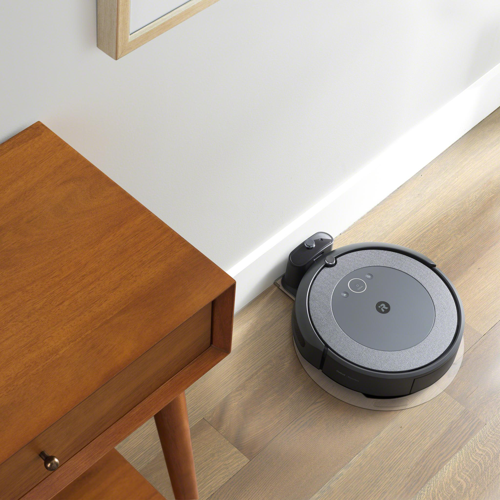Roboter-Staubsauger auf Holzboden, in der Nähe eines Holztisches und einer weißen Wand.