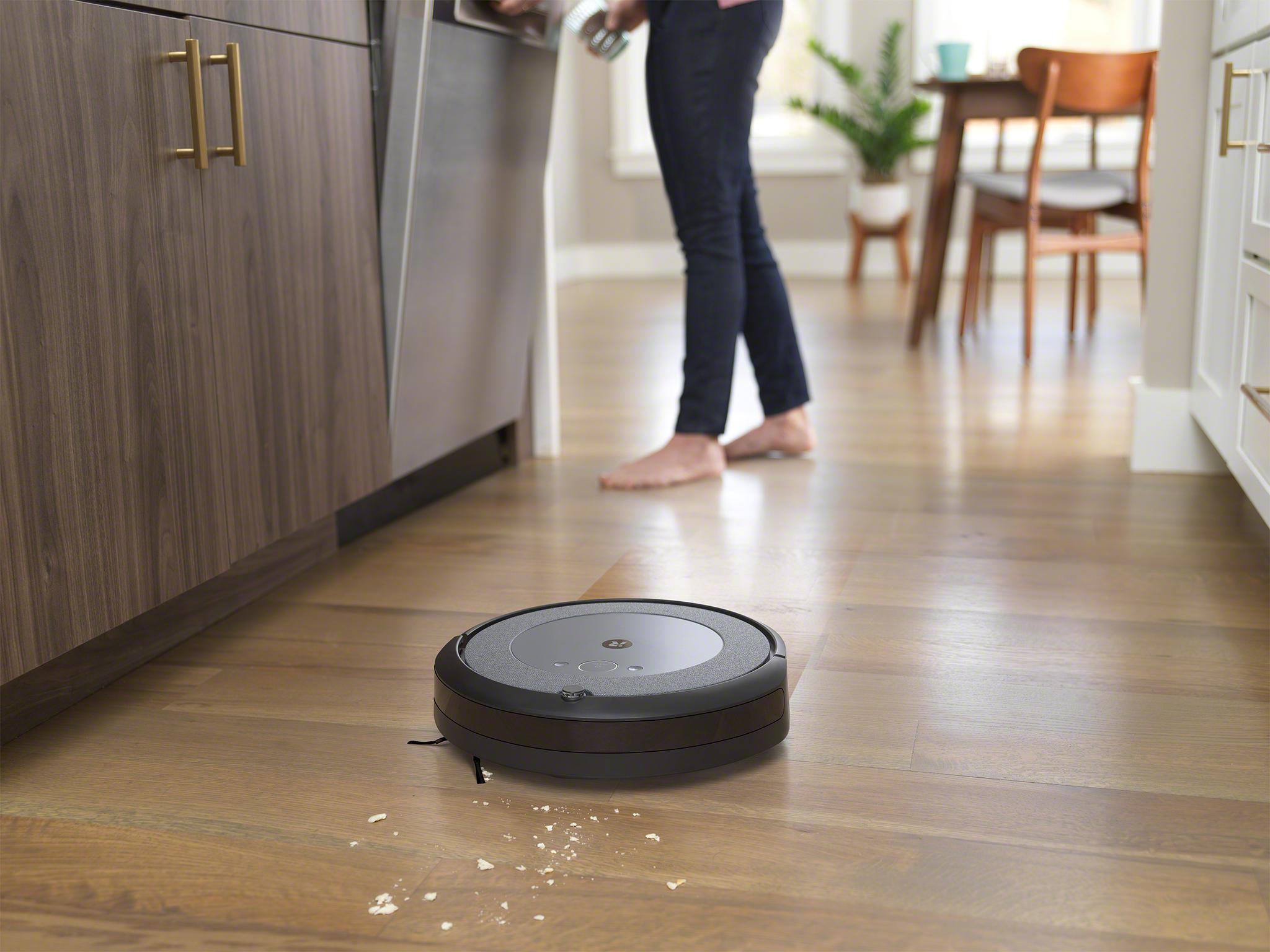 Ein Roboter-Staubsauger auf einem Holzboden, im Hintergrund die Beine einer Person.