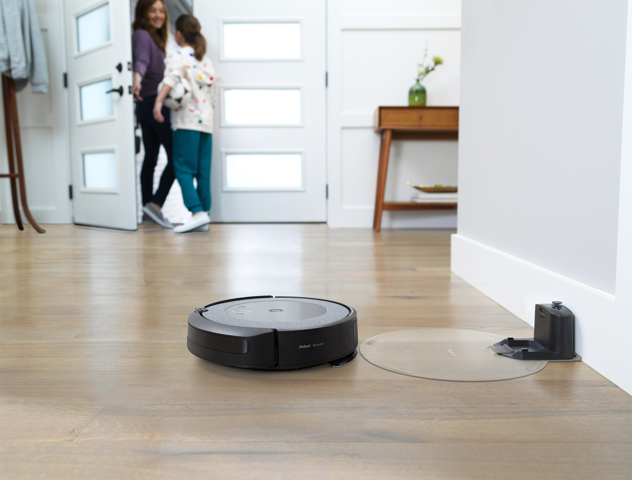 Schwarzer iRobot Roomba Staubsauger auf einem Holzboden. Zwei Personen sind im Hintergrund sichtbar.