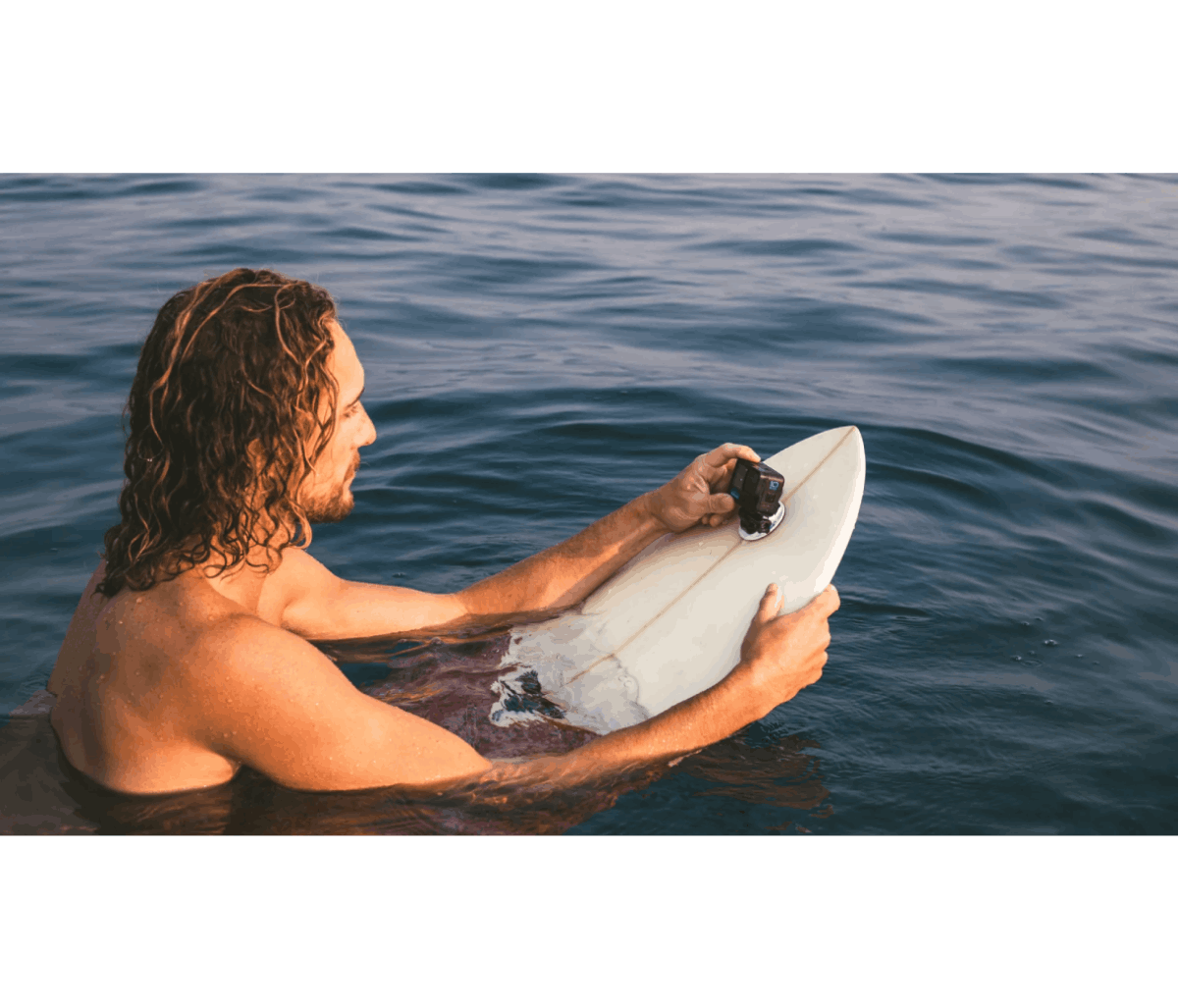 Hombre en una tabla de surf en el agua, sosteniendo una cámara. Está sin camisa. Agua clara y cielo azul.