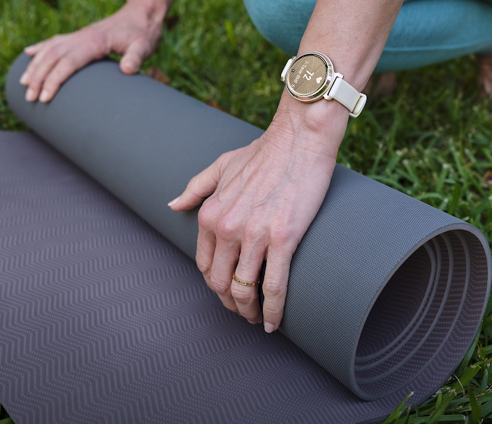Mains enroulant un tapis de yoga gris à l'extérieur sur l'herbe, une montre dorée avec un bracelet blanc est visible.