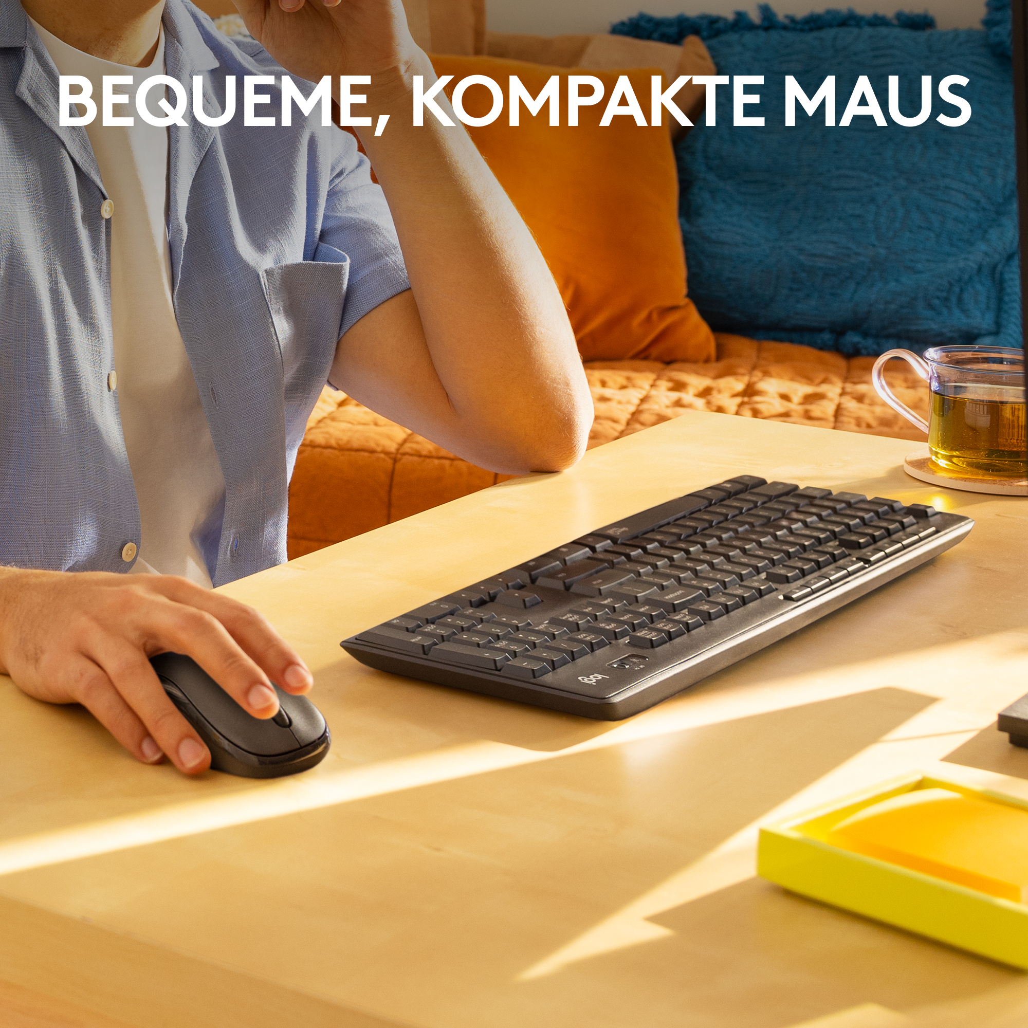 Person benutzt schwarze Maus und Tastatur auf einem Holztisch. Im Hintergrund blaue und orangefarbene Kissen.