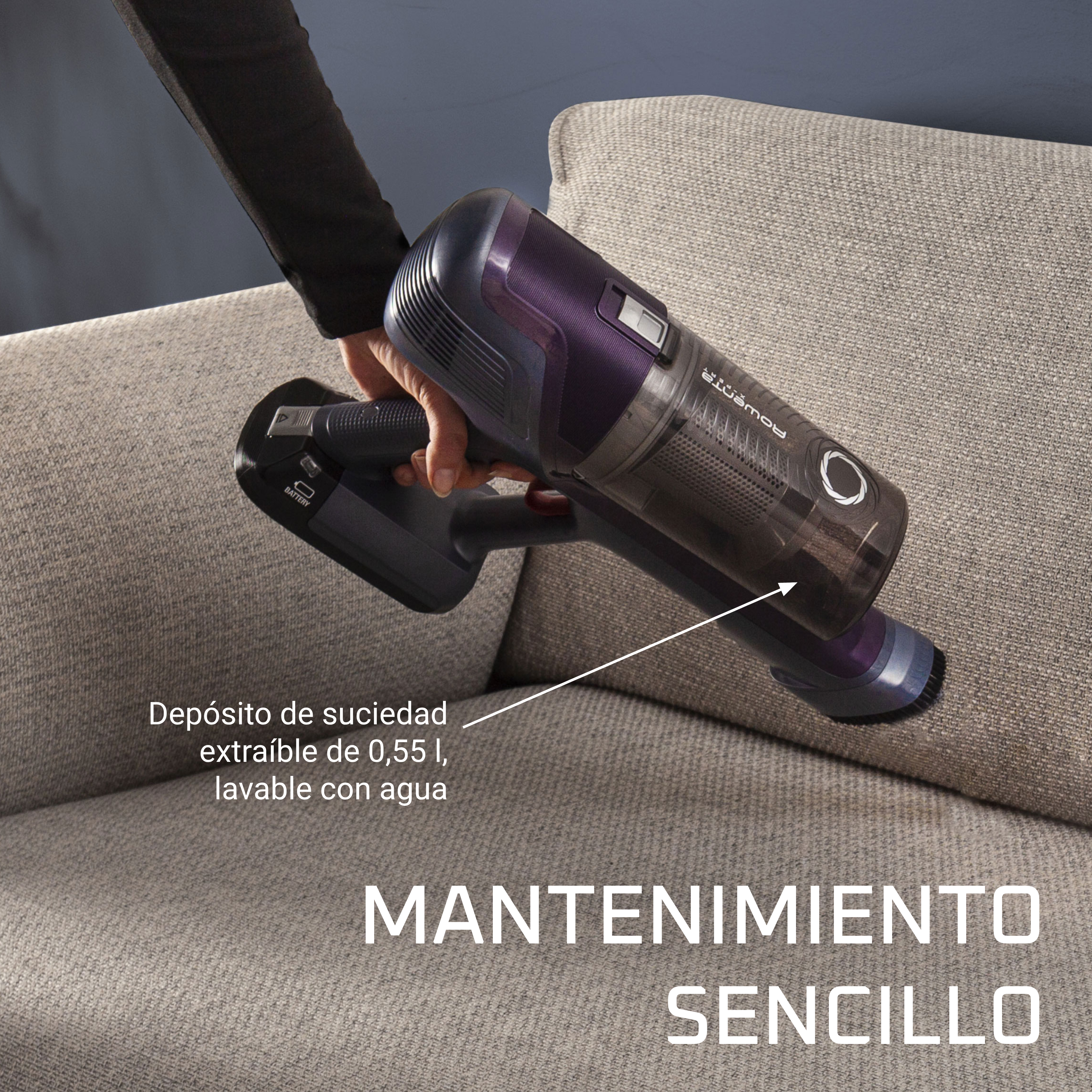 Mano usando una aspiradora de mano en un sofá. La aspiradora es morada oscura y negra.