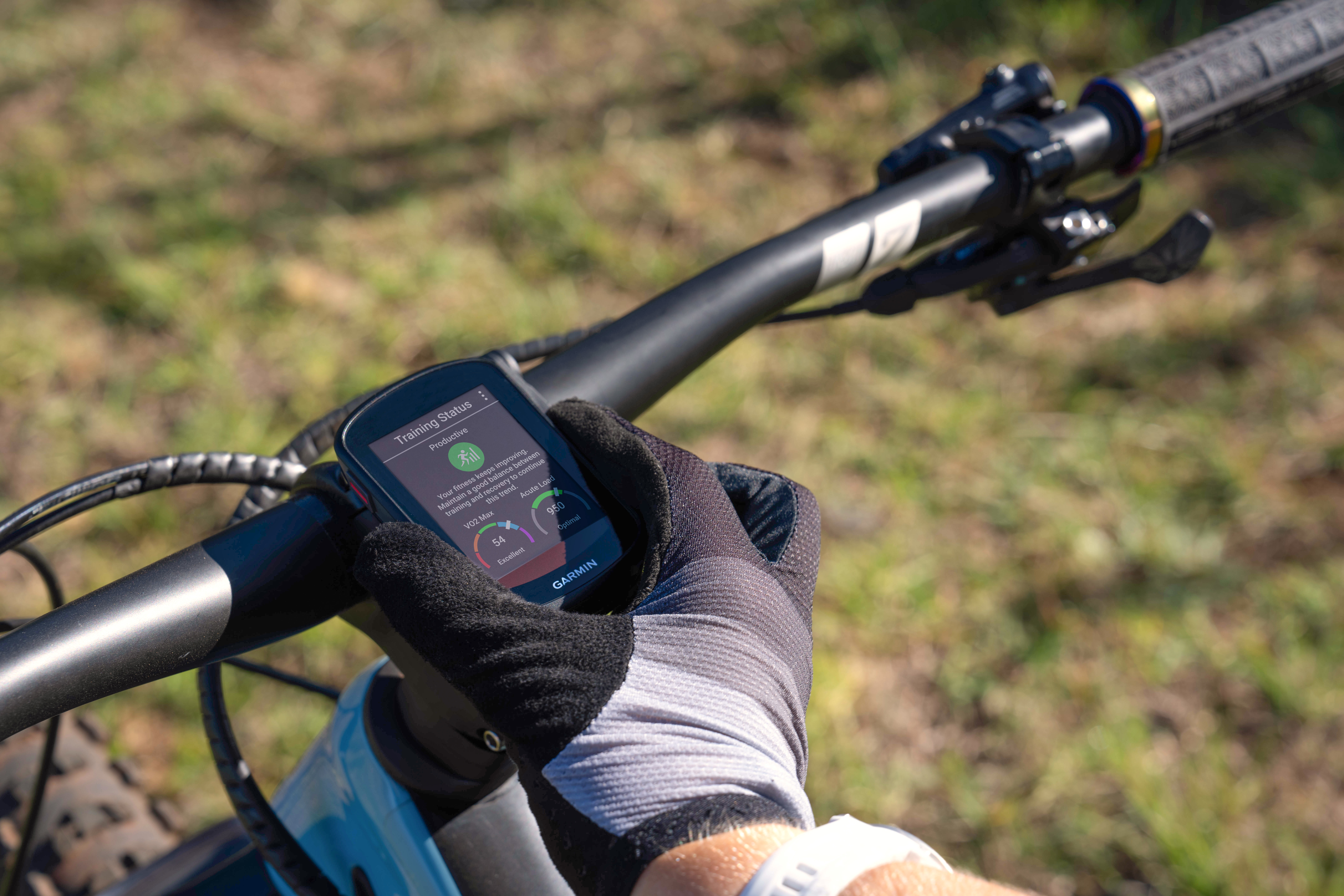 La mano di un ciclista con un guanto tiene un dispositivo Garmin sul manubrio di una mountain bike. Lo sfondo è sfocato.