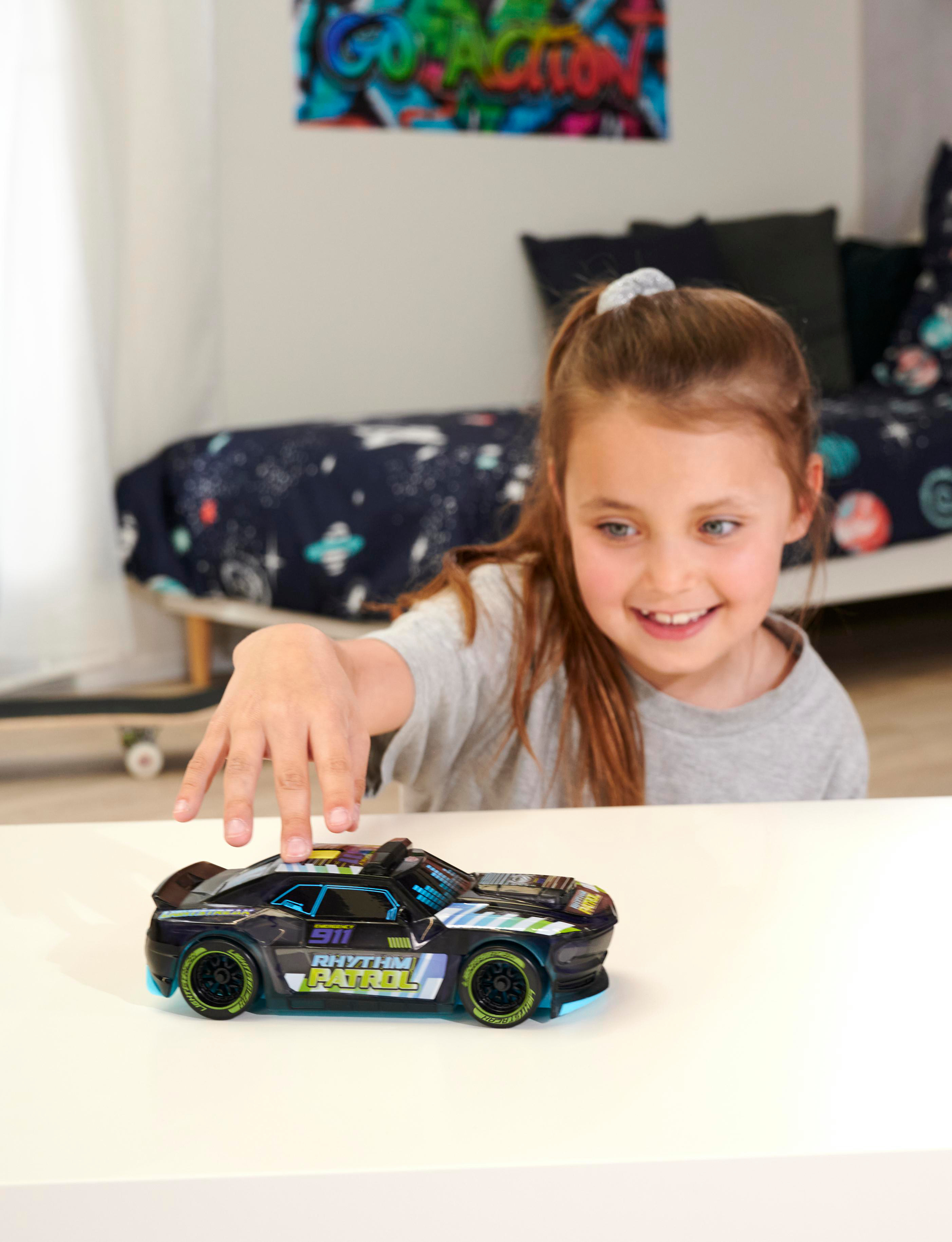 Ein Mädchen greift nach einem Spielzeugauto auf einem weißen Tisch. Ein Bett und Kunstwerk sind im Hintergrund.