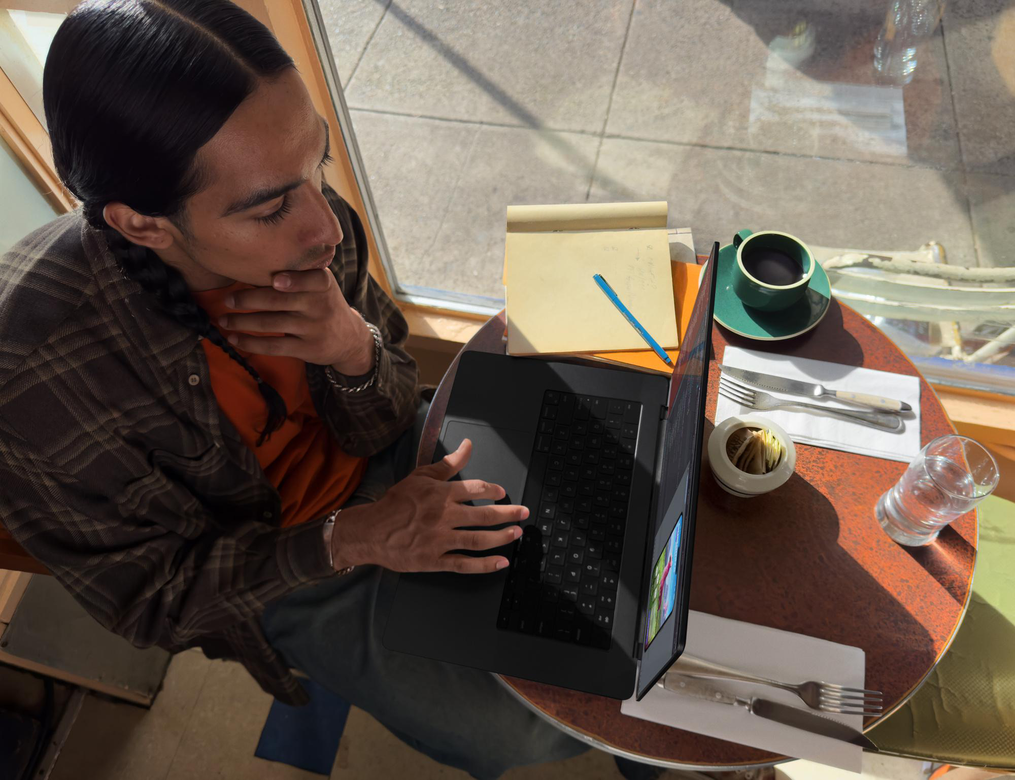 Draufsicht eines Mannes, der an einem Cafétisch mit Notizblock und Kaffee einen Laptop benutzt.