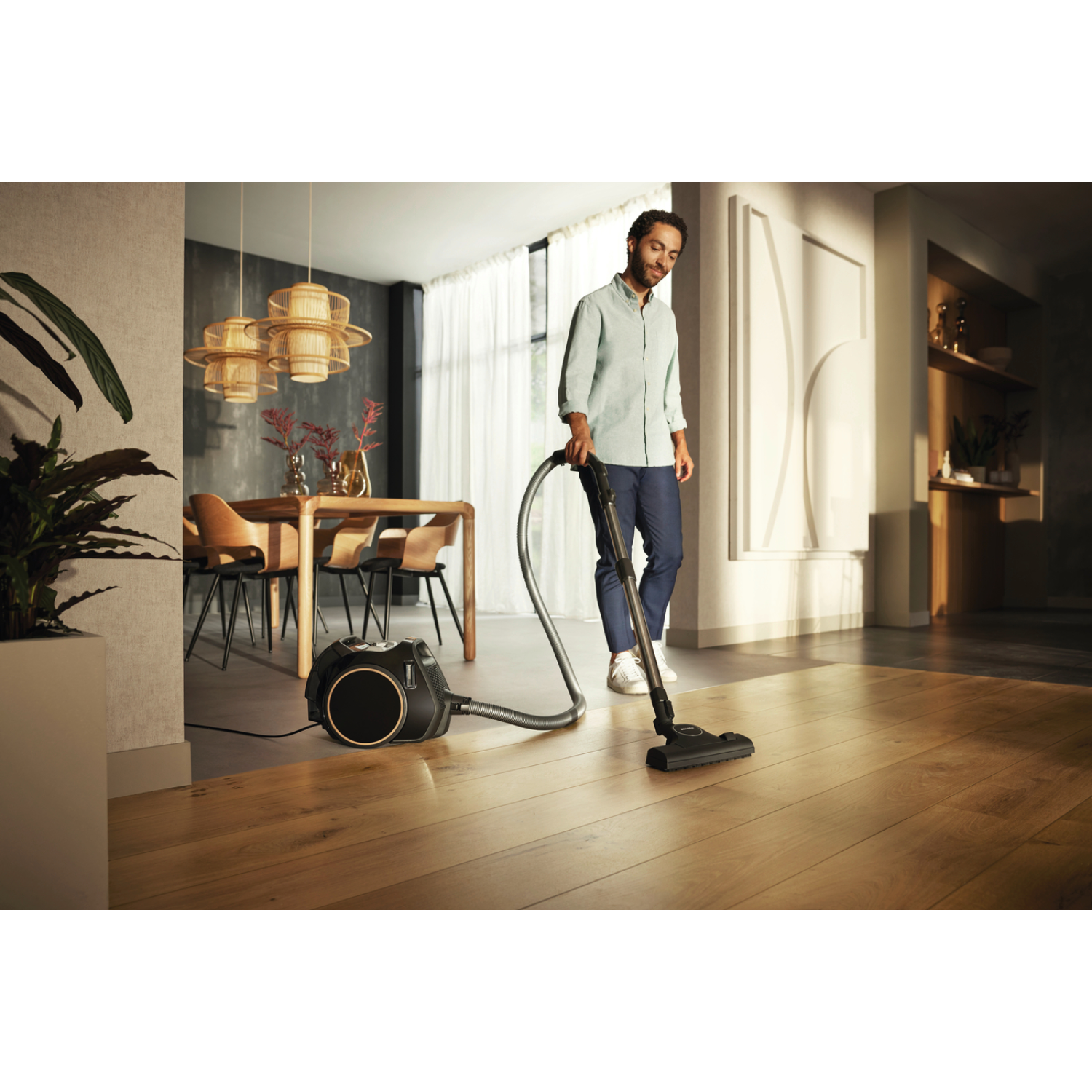 Man vacuums a wood floor. Vacuum cleaner is black. Background: dining table, chairs.