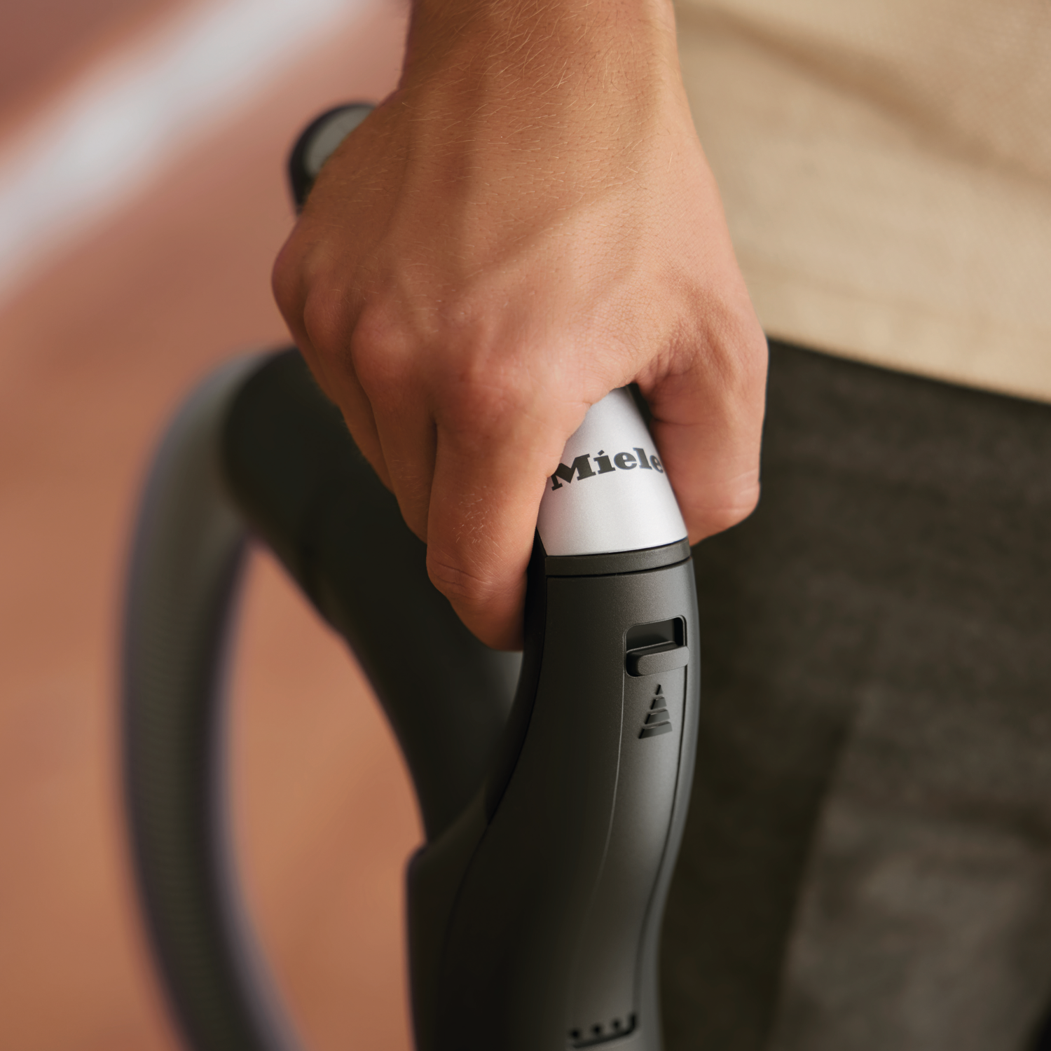 Hand holding a vacuum cleaner handle with the 'Miele' logo. Cream-colored background.