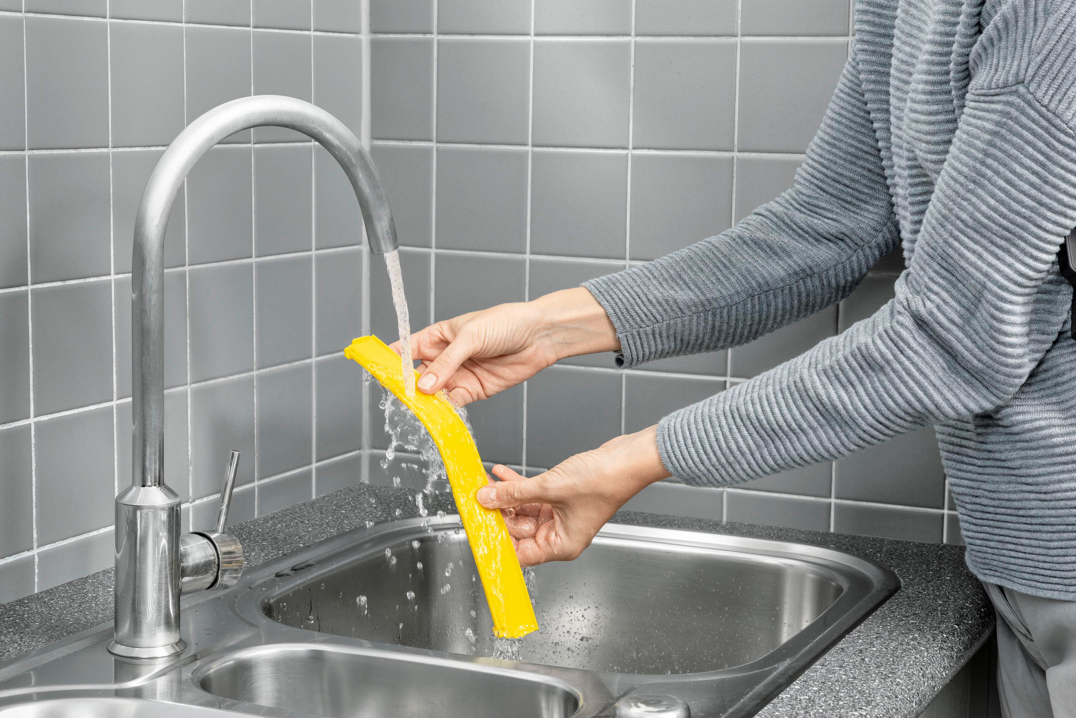 Hände waschen einen gelben nudelartigen Gegenstand unter einem Wasserhahn. Eine grau geflieste Wand ist sichtbar.