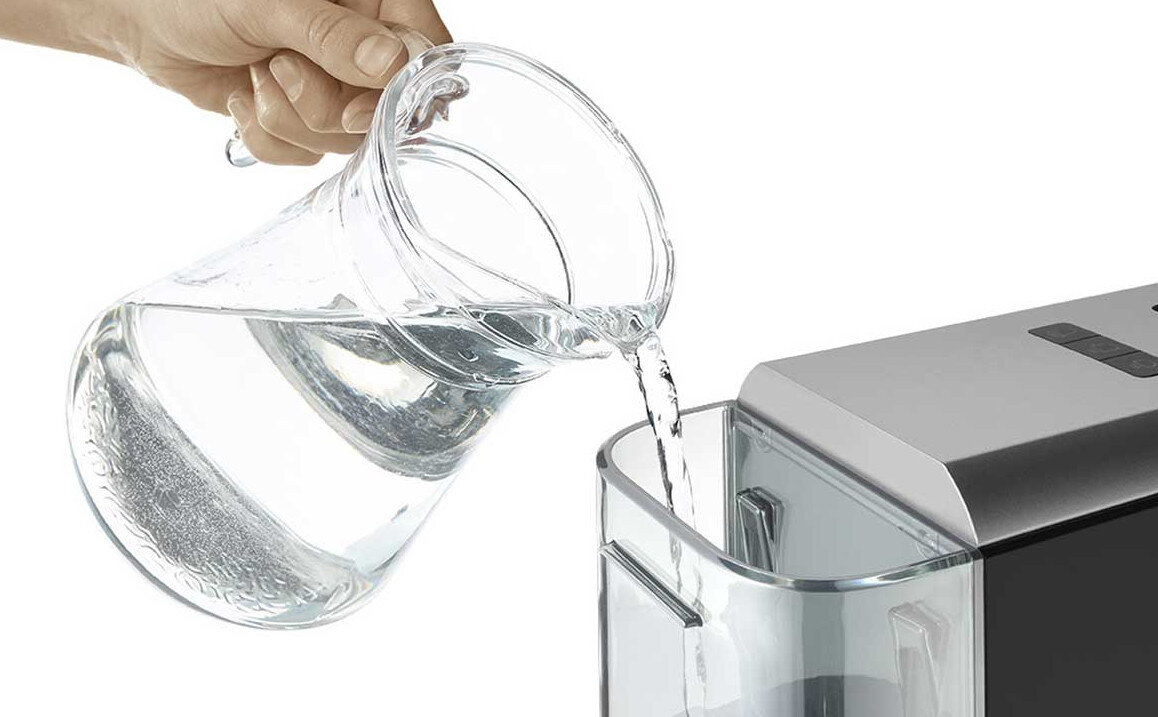 Ręka nalewa wodę ze szklanego dzbanka do przezroczystego pojemnika. Białe tło, zbliżenie.