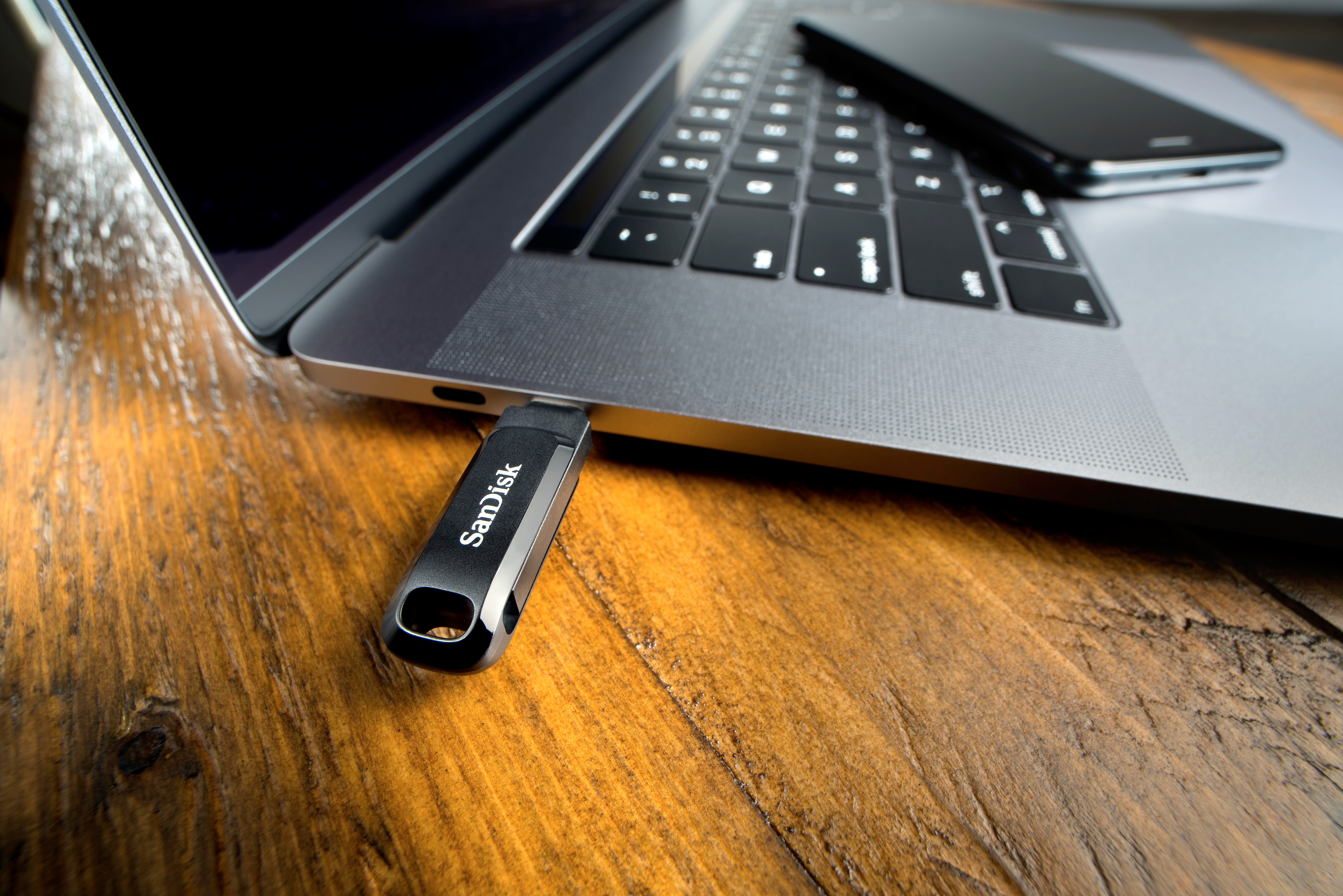 Ein SanDisk USB-Stick in einem Laptop, daneben ein Telefon auf einem Holztisch.