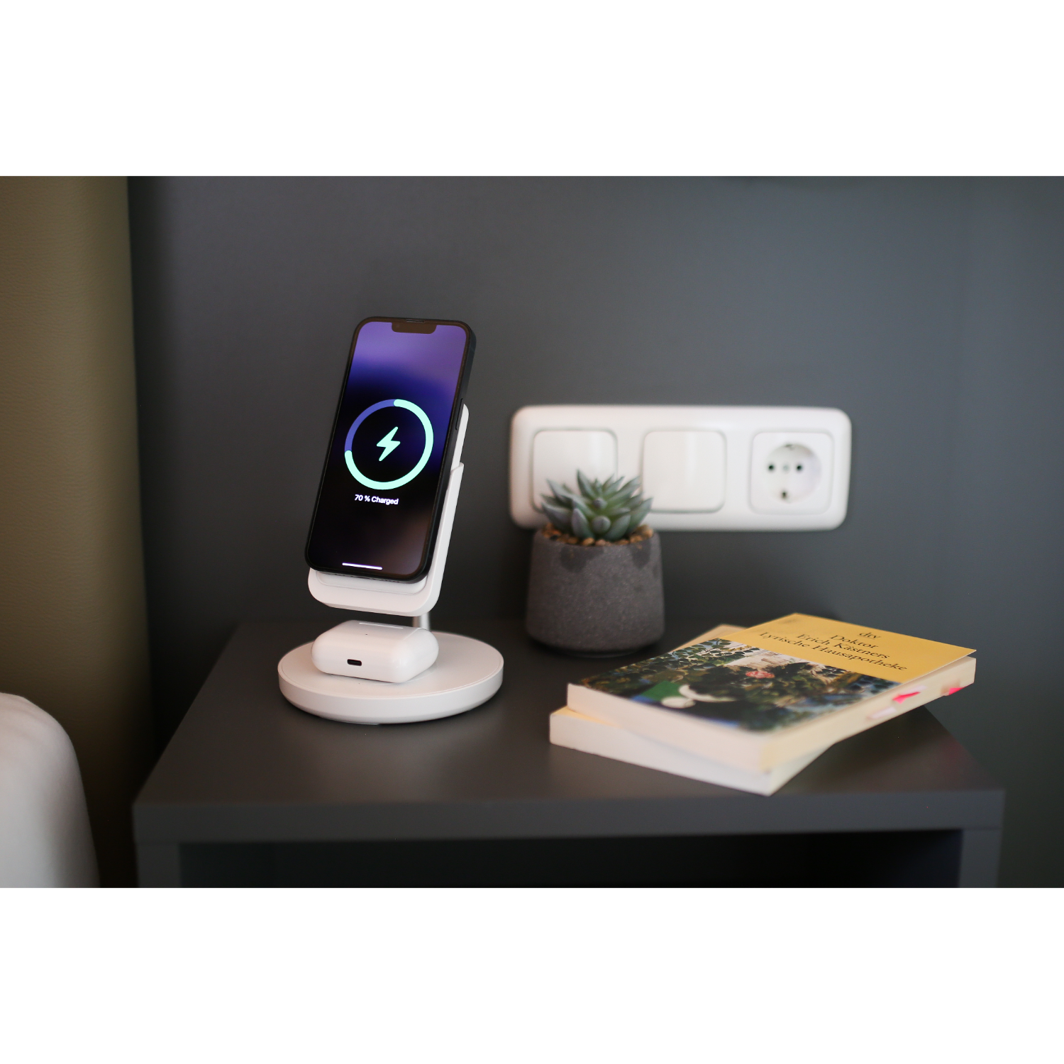 A phone charging on a white stand, book and a small plant on a table, gray wall background.