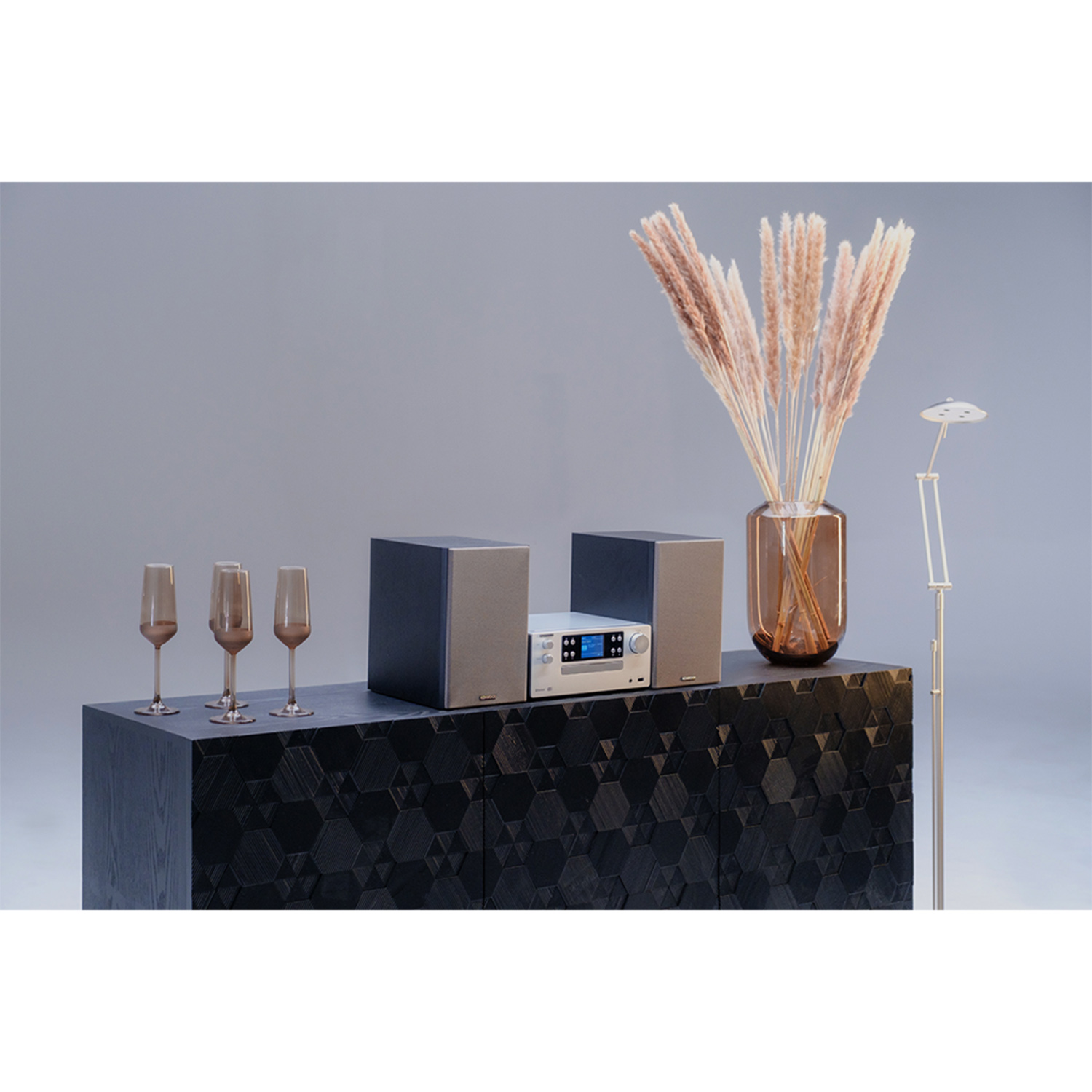 Un mobile in legno scuro con altoparlanti, uno stereo e calici da champagne. Sono presenti fiori secchi e una lampada.
