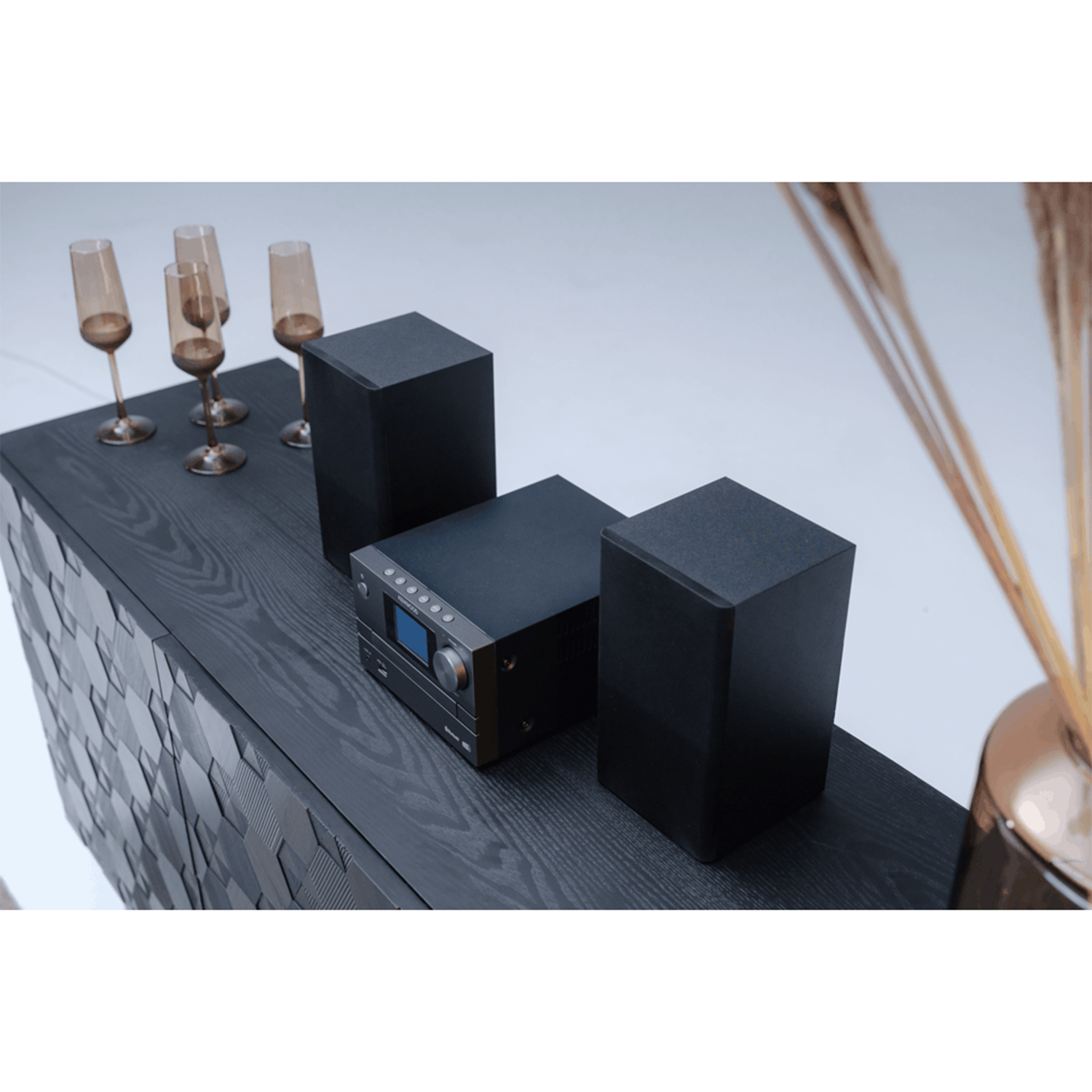 Un impianto stereo nero con due altoparlanti, un ricevitore e quattro bicchieri su un mobile in legno scuro.