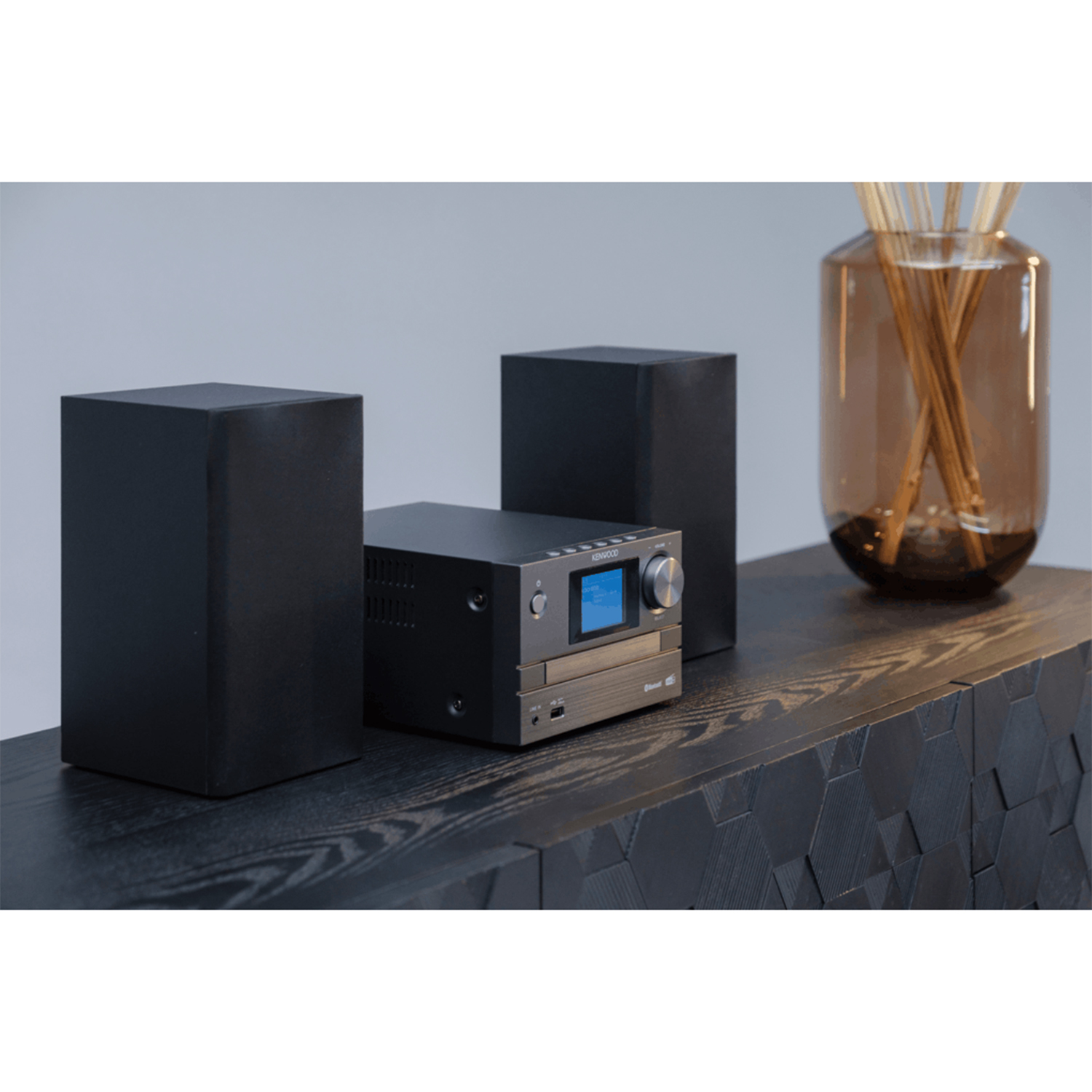 Un impianto stereo nero con altoparlanti su una superficie di legno scuro, con un vaso con bastoncini.