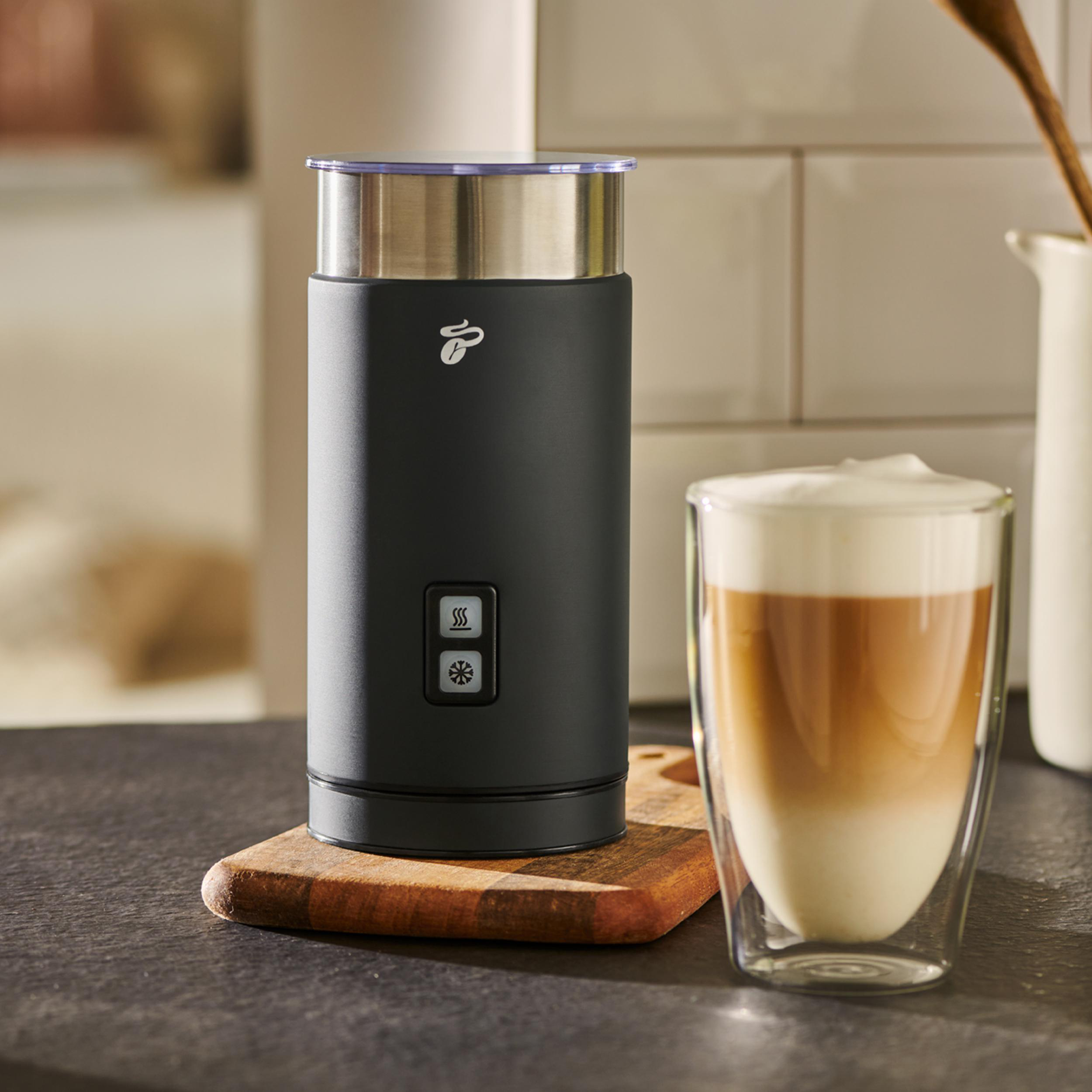 Ein schwarzer Milchaufschäumer auf einem Holzbrett neben einem Glas Latte und einer weißen Kachelwand.