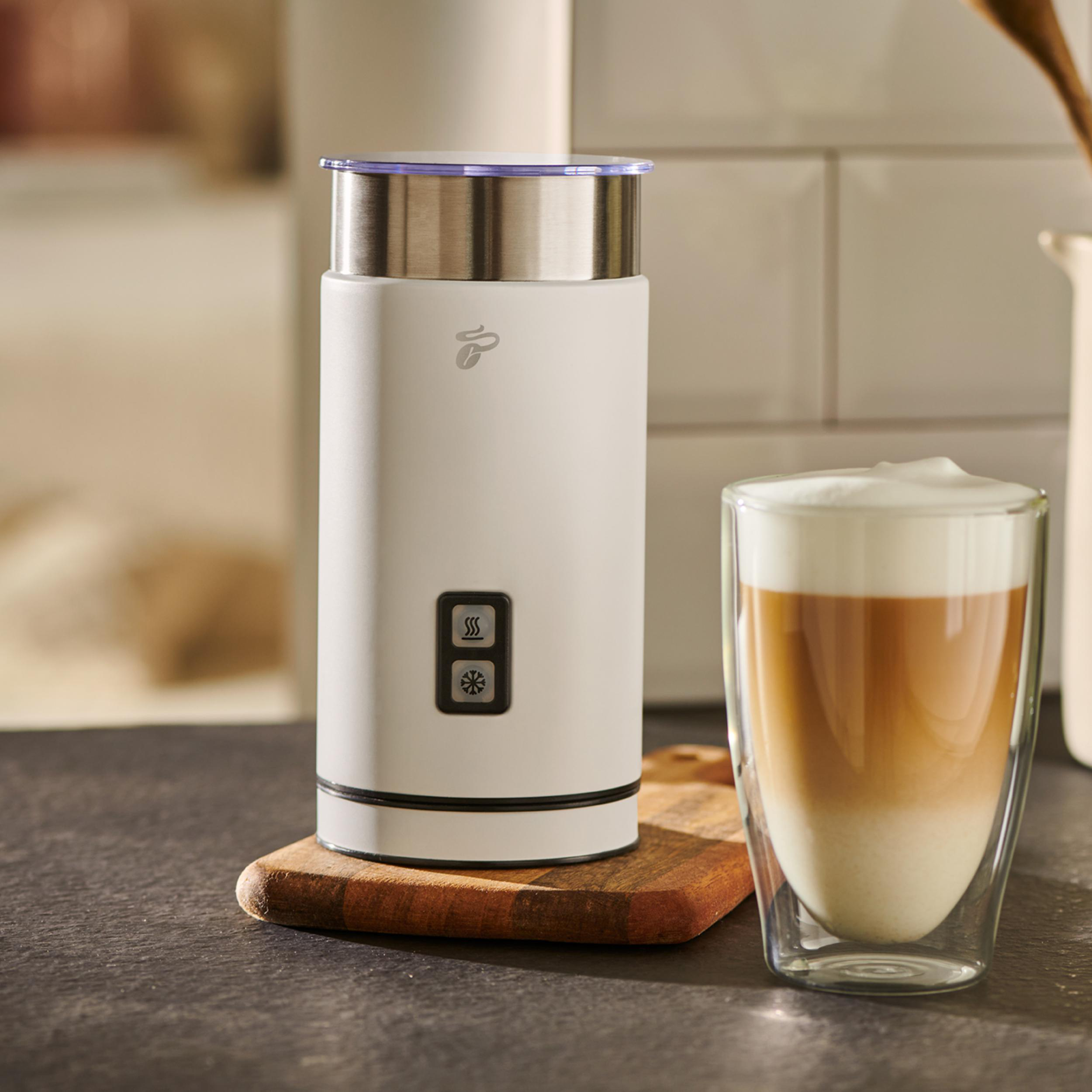 Weißer Milchaufschäumer und ein Glas Latte auf einem Holzbrett. Küchenhintergrund mit Fliesen.