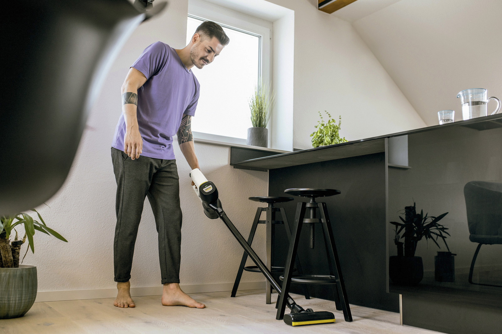 Egy férfi egy konyhát porszívózik egy vezeték nélküli porszívóval. Lila pólót és szürke nadrágot visel.