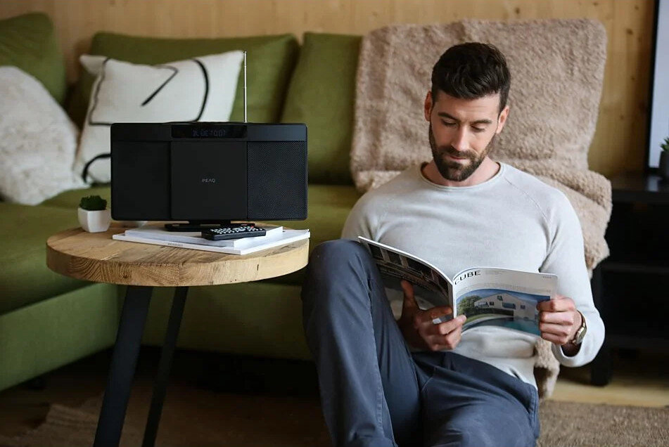 Mężczyzna czyta magazyn, siedząc obok systemu głośnikowego na drewnianym stoliku.