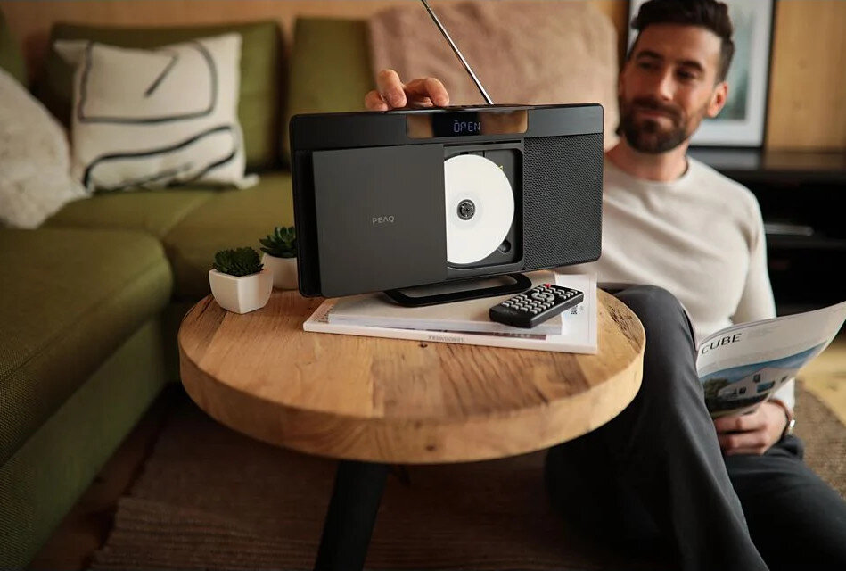 Mężczyzna siedzi z odtwarzaczem CD na małym stoliku, czytając czasopismo. W tle sofa i rośliny.