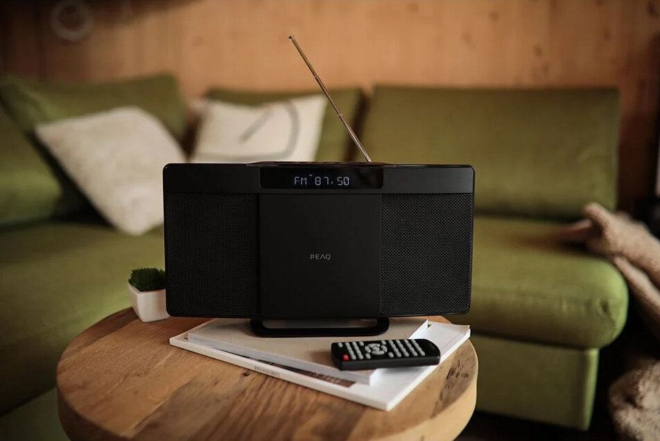 Czarny radioodtwarzacz na drewnianym stoliku, przed zieloną sofą. Na stole również pilot i książki.