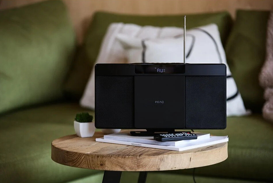 Czarny głośnik na drewnianym stole z pilotem. Zielona sofa w tle.