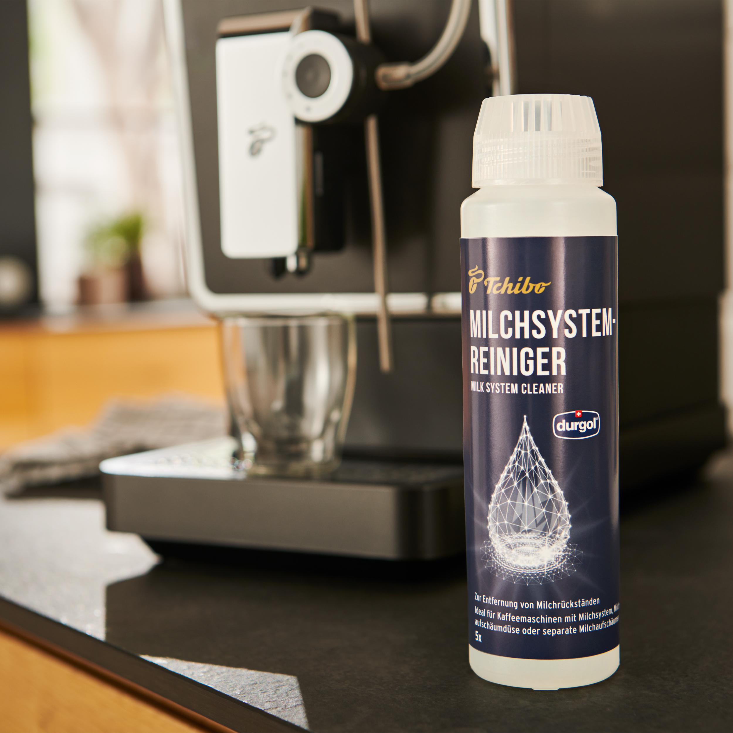 Eine Flasche Milchsystemreiniger neben einer Kaffeemaschine, mit einem Glas im Hintergrund.