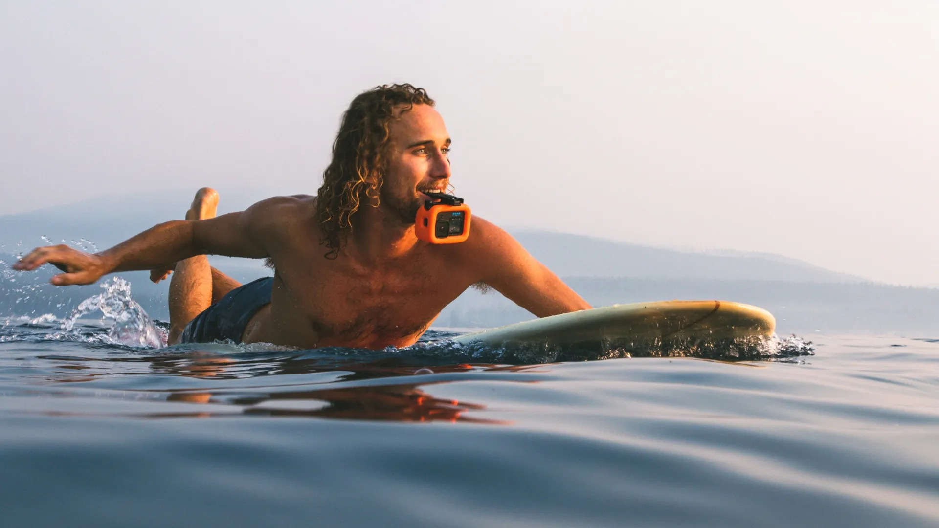 Ein Mann auf einem Surfbrett liegt auf dem Bauch, mit einer orangefarbenen Kamera im Mund, im Meer.