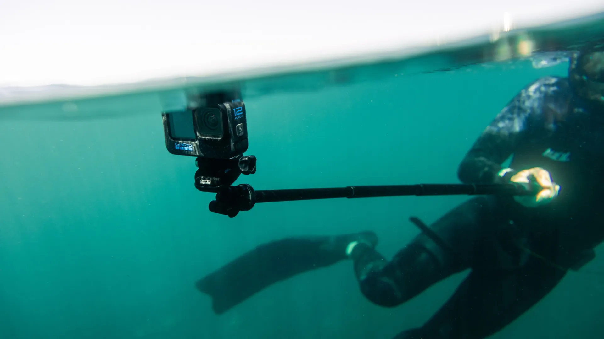 Eine GoPro 12 Black Kamera an einem Stab, teilweise untergetaucht in türkisblauem Wasser.