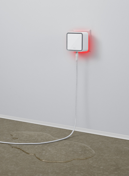 Ein weißes Smart-Home-Gerät in der Wand, rotes Licht. Wasserpfütze auf dem Boden.