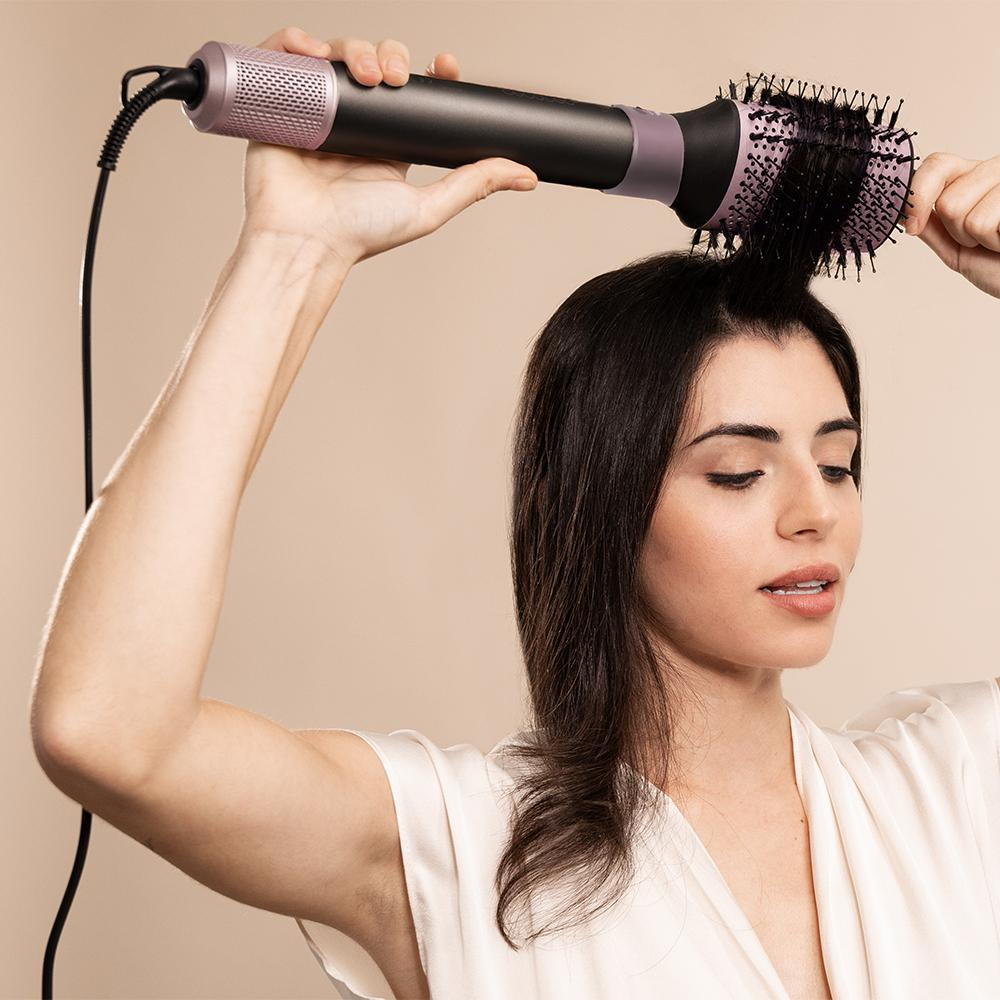 Mujer peinando su cabello oscuro con un cepillo secador de pelo negro y lavanda. Lleva una bata blanca.