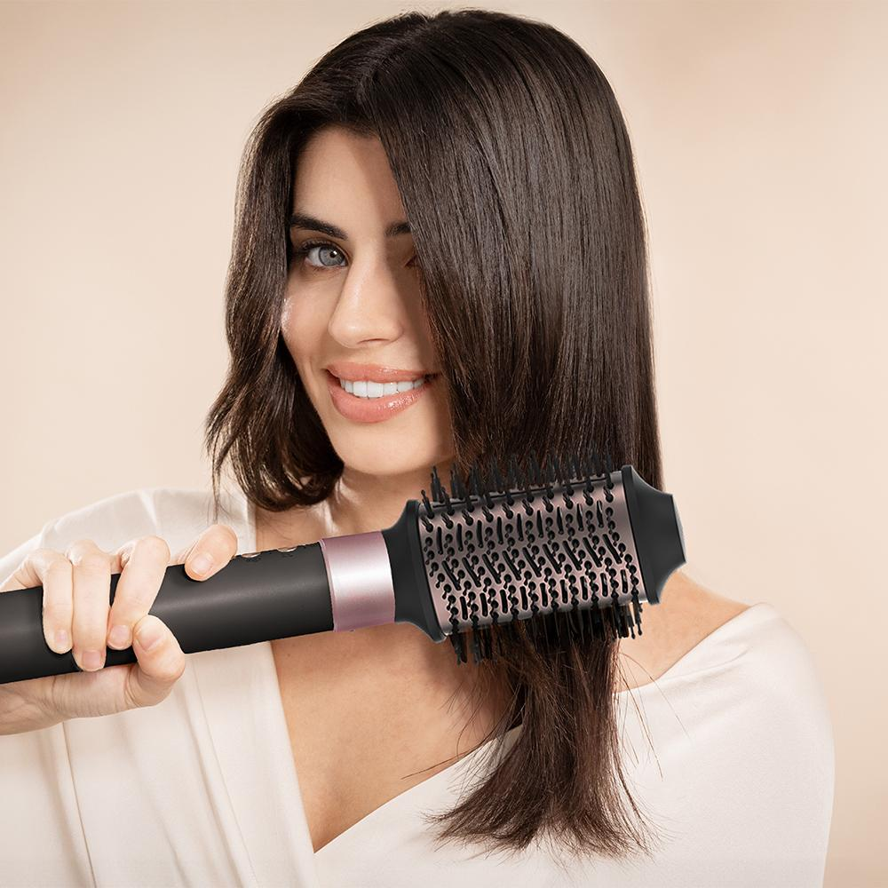 Una mujer cepillando su cabello con un cepillo redondo, sonriendo a la cámara.