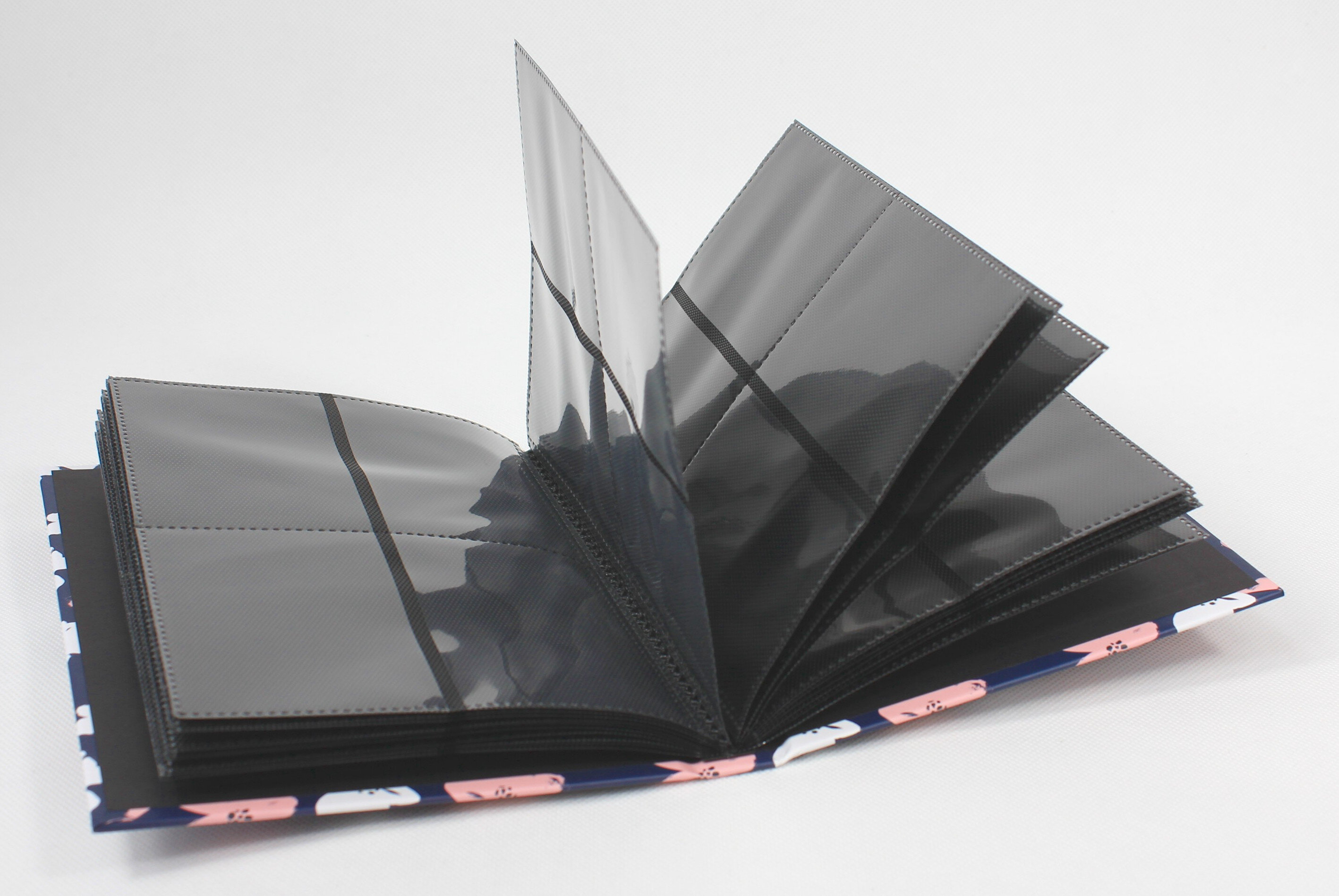 Otwarty album fotograficzny ze stronami i kieszeniami. Niebieska okładka z białym i różowym wzorem kwiatowym. Białe tło.