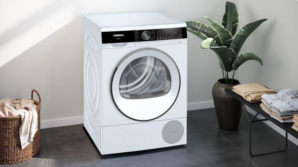 White dryer. Plant and clothes in the background. White floor and wall.