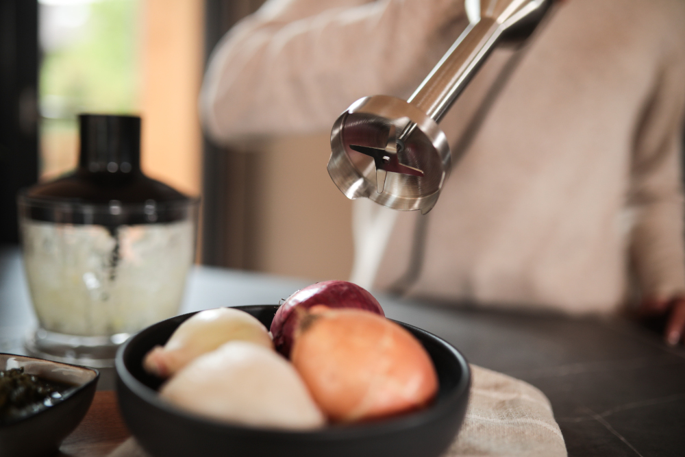 A blender head is above a bowl of onions, with a person blurred in background.