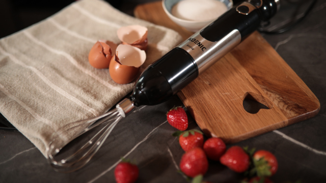 A black and silver hand blender next to a wooden board, eggs, and strawberries.