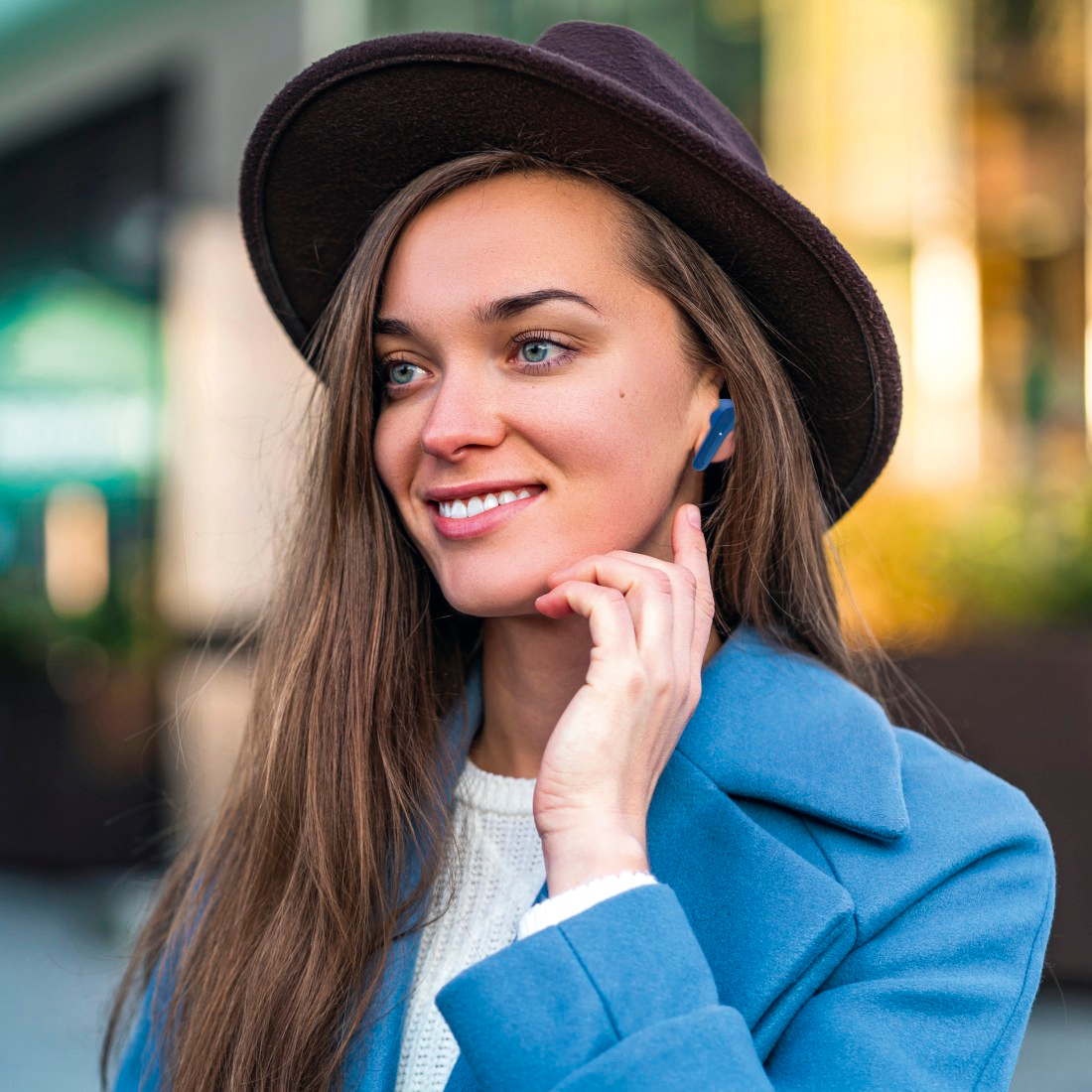 Une femme portant un chapeau et des écouteurs sourit, manteau bleu, en extérieur.