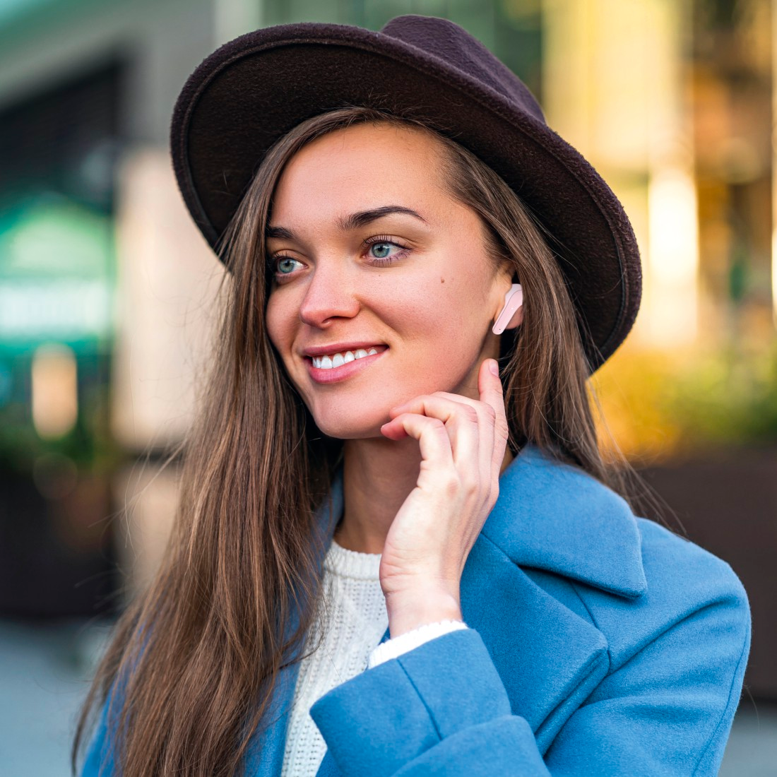Une femme portant un chapeau sourit tout en portant des écouteurs roses. Elle a les cheveux longs et est à l'extérieur.