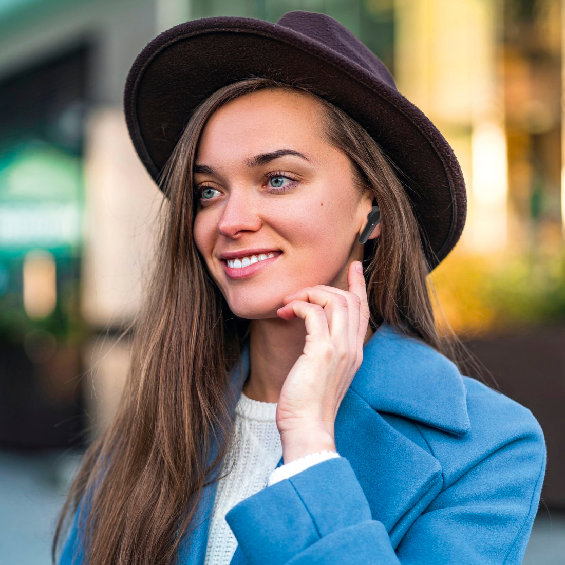 Femme avec un chapeau et un manteau bleu, portant un écouteur, souriante, en extérieur.