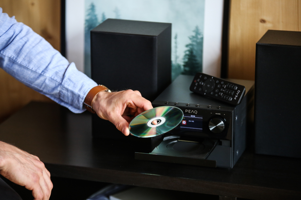 Persoon steekt een cd in een stereosysteem. Zwarte componenten, houten achtergrond.