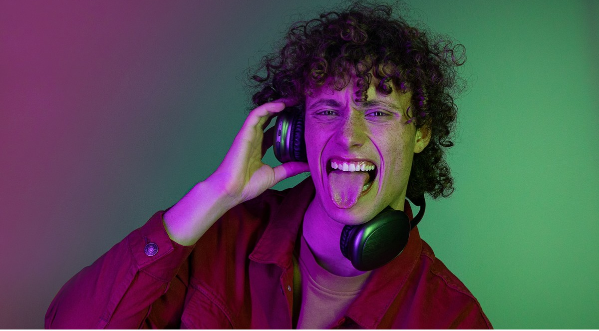 Mann mit Locken, Kopfhörern, Zunge herausstrecken, rotes Hemd, grüner und pinker Hintergrund.