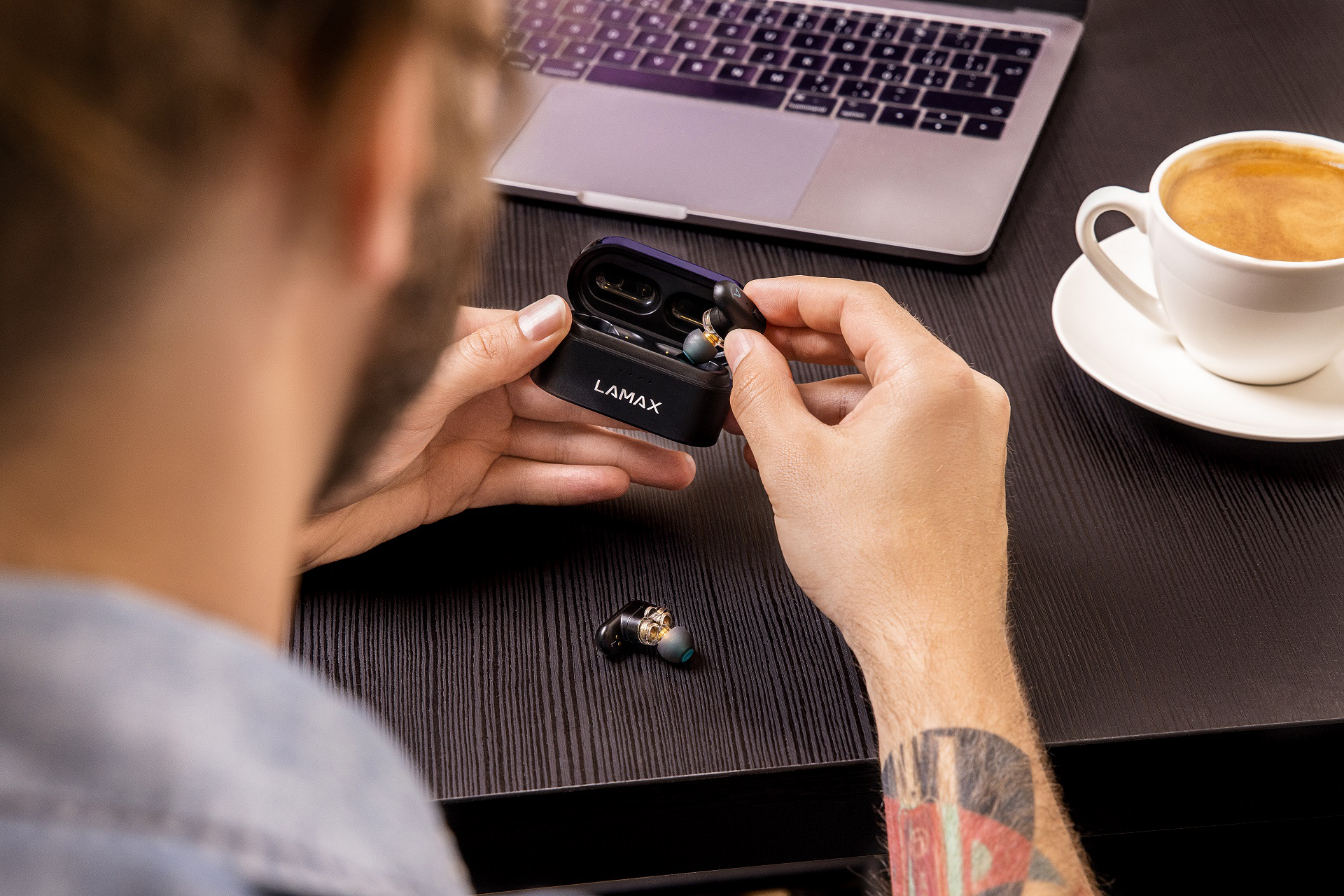 Személy fekete LAMAX fülhallgató tokot és egy fülhallgatót tart. Laptop és kávéscsésze.