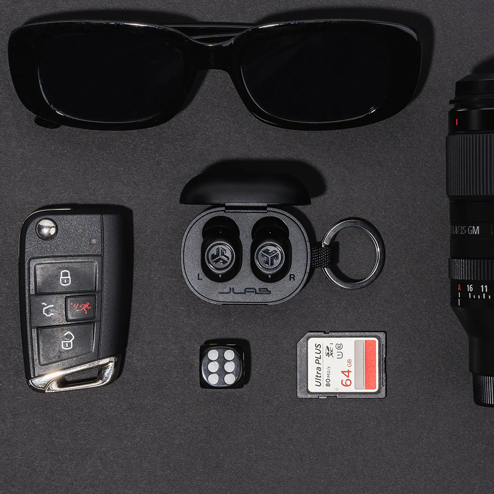 Divers objets sur une surface noire : lunettes de soleil, écouteurs, clé de voiture, dés, carte SD et objectif.