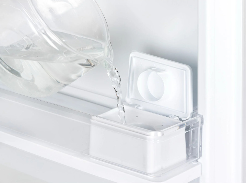 Clear pitcher pouring water into a white container in a refrigerator.