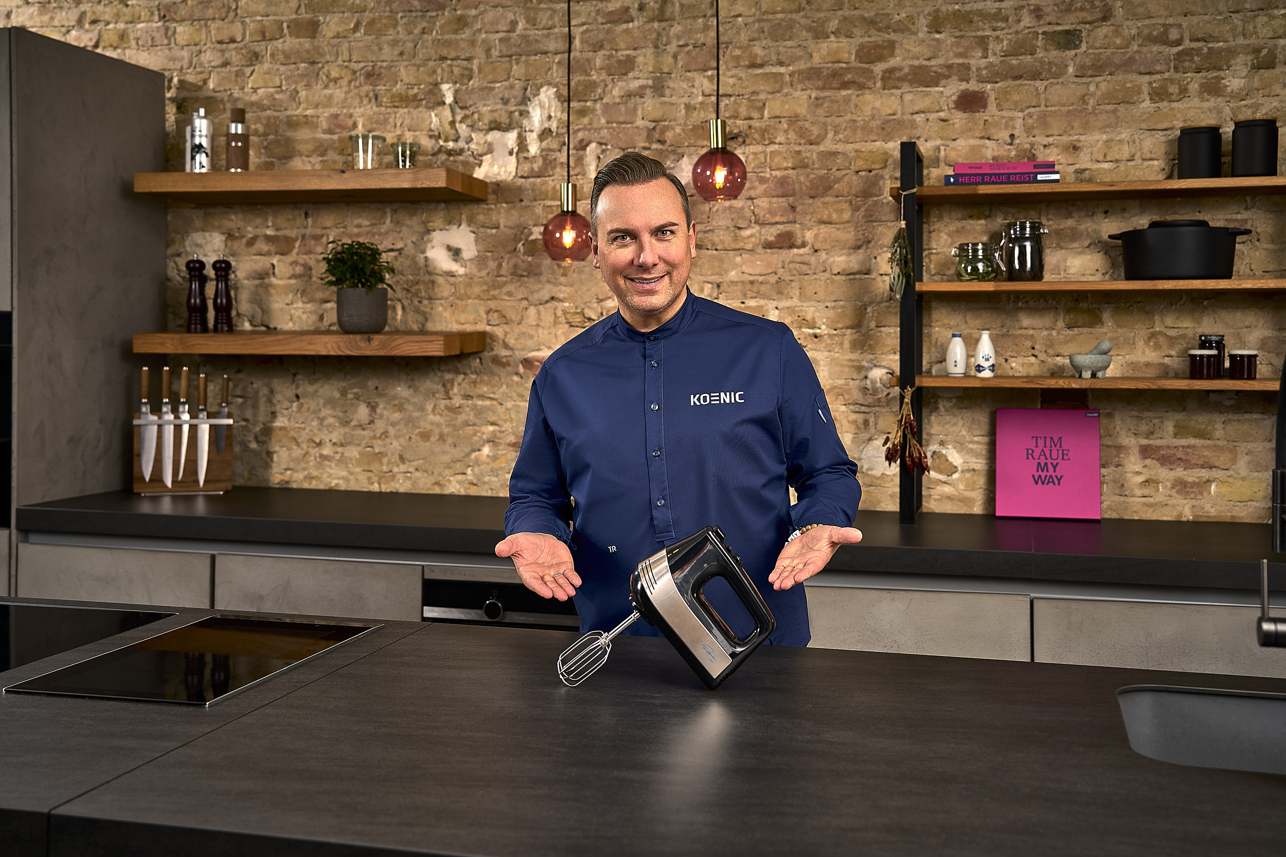Mann mit einer Rührmaschine in einer Küche. Er lächelt und präsentiert die Maschine. Hintergrund ist Küche.
