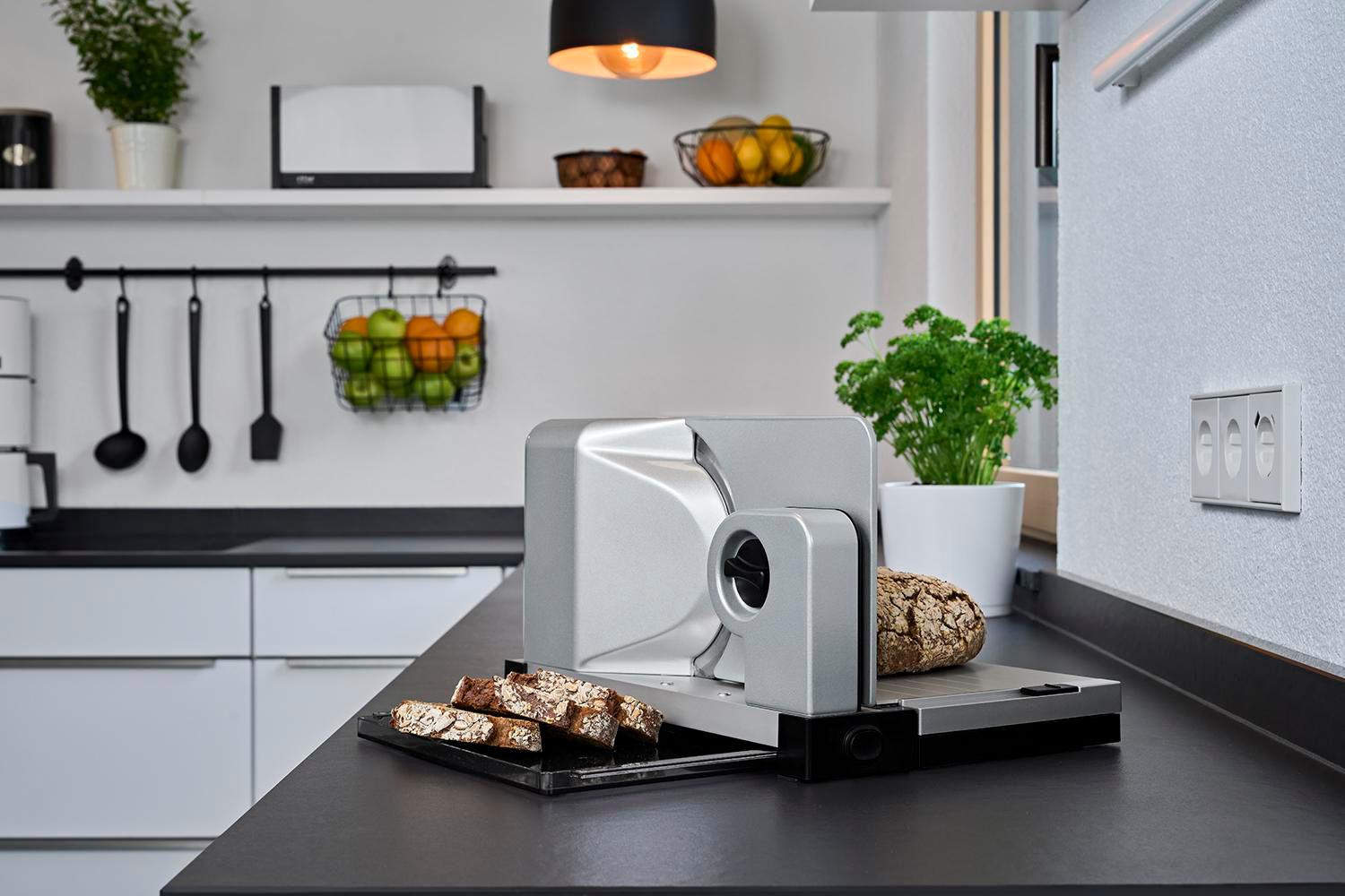 Küchenszene mit Brotschneidemaschine und geschnittenem Brot auf schwarzer Arbeitsplatte.