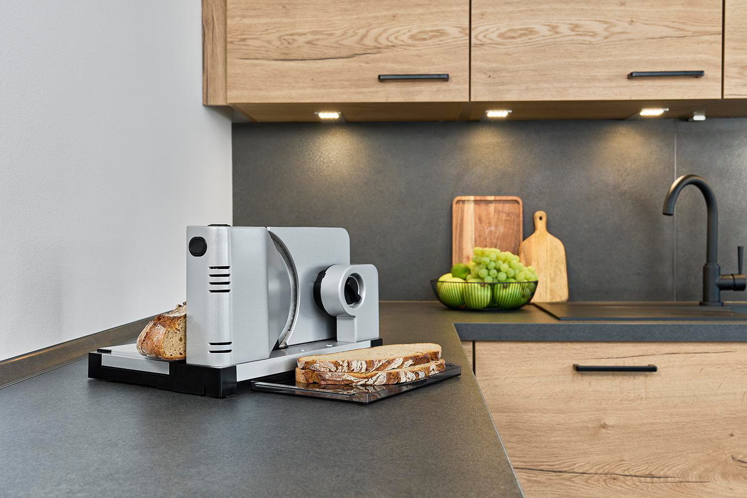 Aufschnittmaschine auf einer Arbeitsplatte, mit Brot und einem Messer. Küchenumgebung mit Holzschränken.